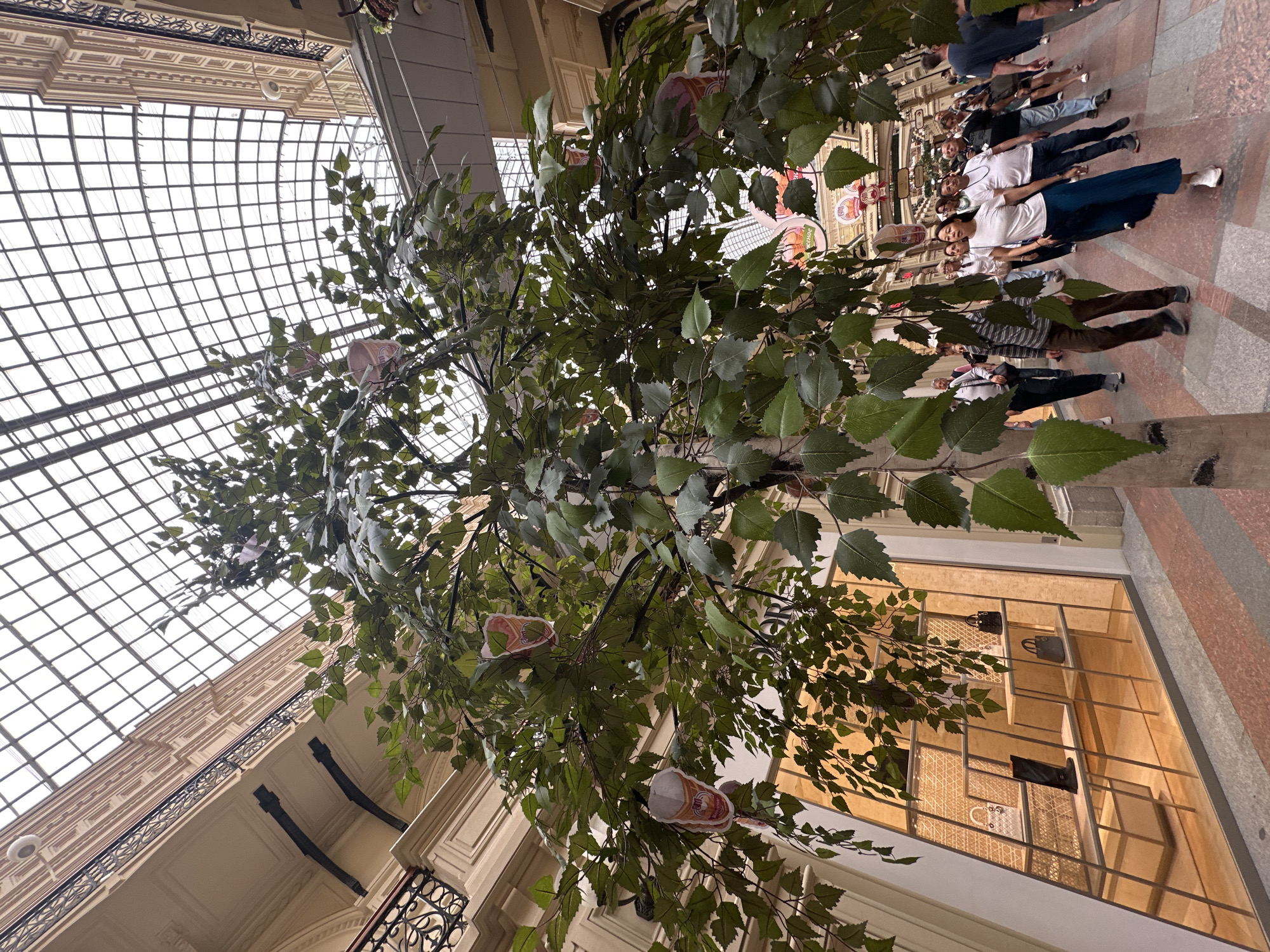 The image depicts the interior of GUM department store on Red Square in Moscow, Russia. A large, artificial tree with green leaves is prominently displayed in the foreground, with some decorative elements resembling ice cream cones hanging from its branches. People are seen walking through the spacious, multi-level interior, which features a distinctive glass-paneled ceiling and ornate architectural details. The flooring is a patterned marble. Shops with illuminated displays of merchandise, including handbags and shoes, are visible along the sides of the walkway. The overall atmosphere appears to be that of a busy, upscale shopping environment.