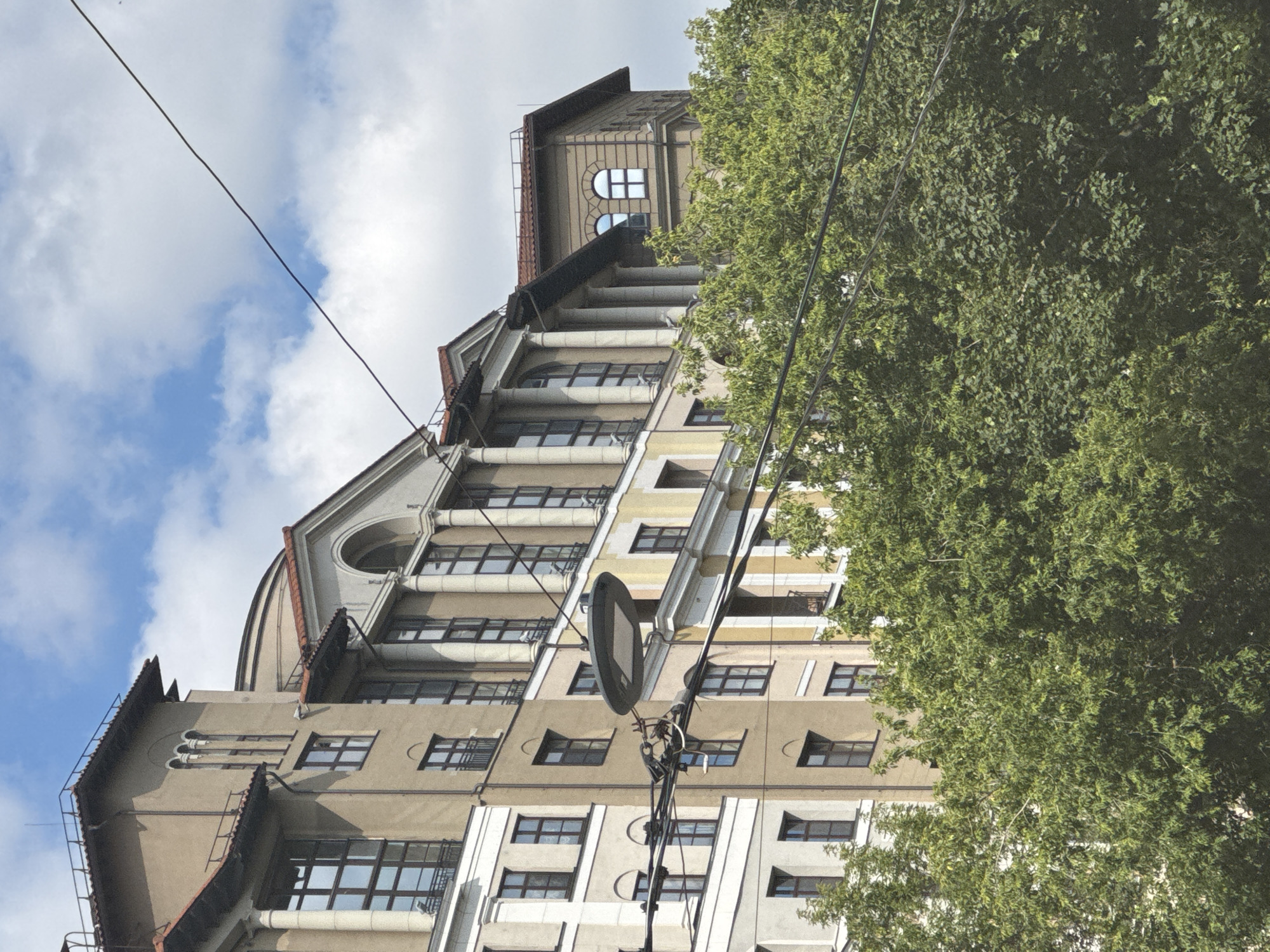 This is a low-angle, vertical photograph of a grand, multi-story building with classical architectural elements, possibly from the early 20th century. The building features a light beige facade, ornate cornices, arched windows, and balconies. Several windows are visible on different levels, many with dark frames and a grid-like structure. The roofline is complex, with several gabled sections and a prominent dormer window at the top.
A large, leafy green tree dominates the right side of the frame, obscuring parts of the building and the sky. The tree appears full and healthy, suggesting it is springtime or summer. In the background, a cloudy sky with patches of blue is visible.
Two dark wires run diagonally across the sky and down the side of the building, suggesting the presence of overhead electrical or telecommunication lines. A circular, dark-colored street lamp or sign fixture is attached to a pole with wires, positioned in front of the building.
The lighting suggests it is daytime, likely midday, with bright but diffused sunlight due to the clouds. The scene conveys a sense of urban architecture and the integration of nature within a city. There is no visible text in the image. The setting appears to be a street in Moscow, Russia, given the architectural style and urban context. There are no people visible in the photograph.