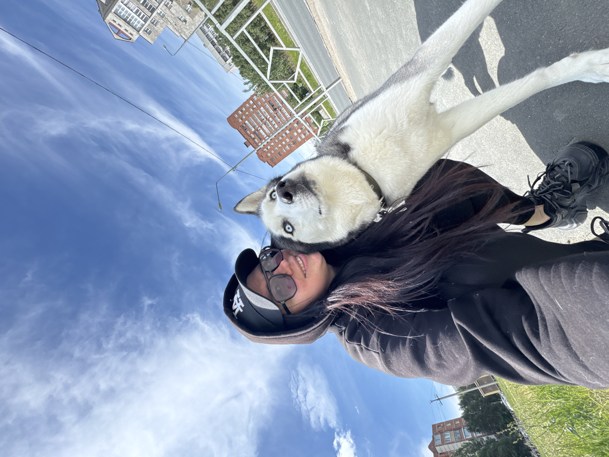 A woman poses for a selfie with a Siberian Husky in an urban setting. The woman, wearing a black hoodie, baseball cap, and sunglasses, smiles at the camera. The Husky, with its white and grey fur and striking blue eyes, rests its head on her shoulder. A street and some buildings are in the background. The sky is blue with scattered clouds.