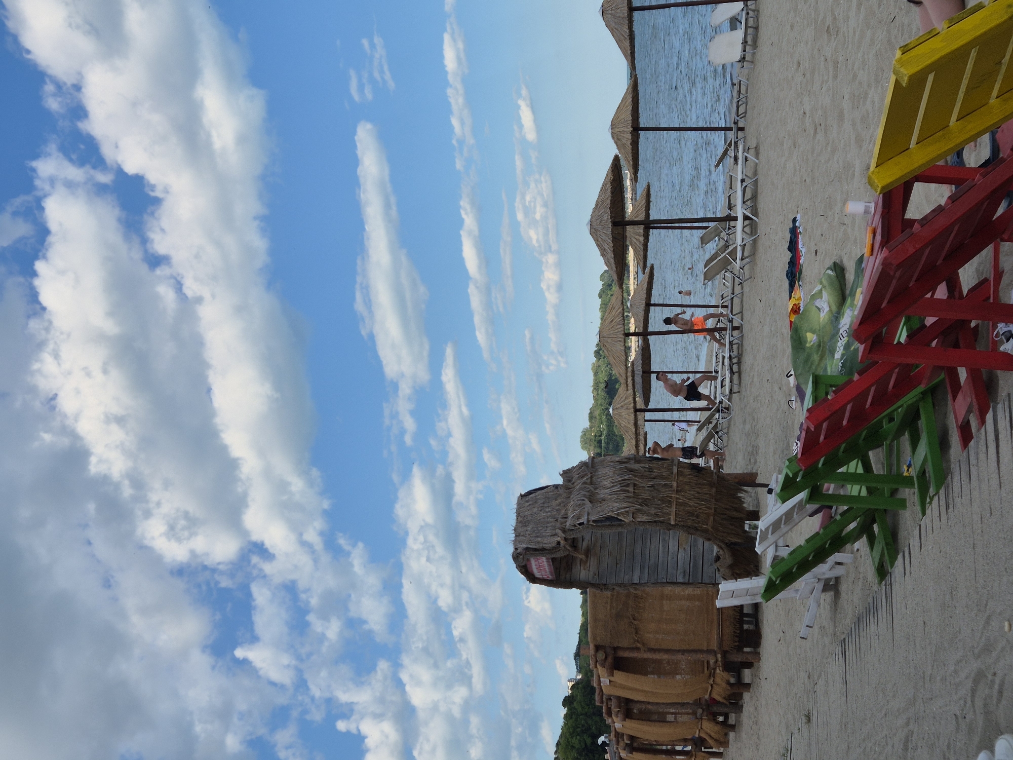 The image depicts a sunny day at a beach, likely in Varna, Bulgaria, with a clear blue sky dotted with white clouds. In the foreground, a wooden walkway leads to a collection of colorful beach chairs, some of which are red, green, and yellow. Beyond the chairs, the sandy beach stretches towards the sea, where several straw umbrellas provide shade. In the middle ground, a rustic wooden structure with a thatched roof stands on the sand, with some cabins or huts visible to its left. People are seen in the water and on the beach, enjoying the seaside atmosphere. The overall scene conveys a relaxed and vibrant beach environment.
