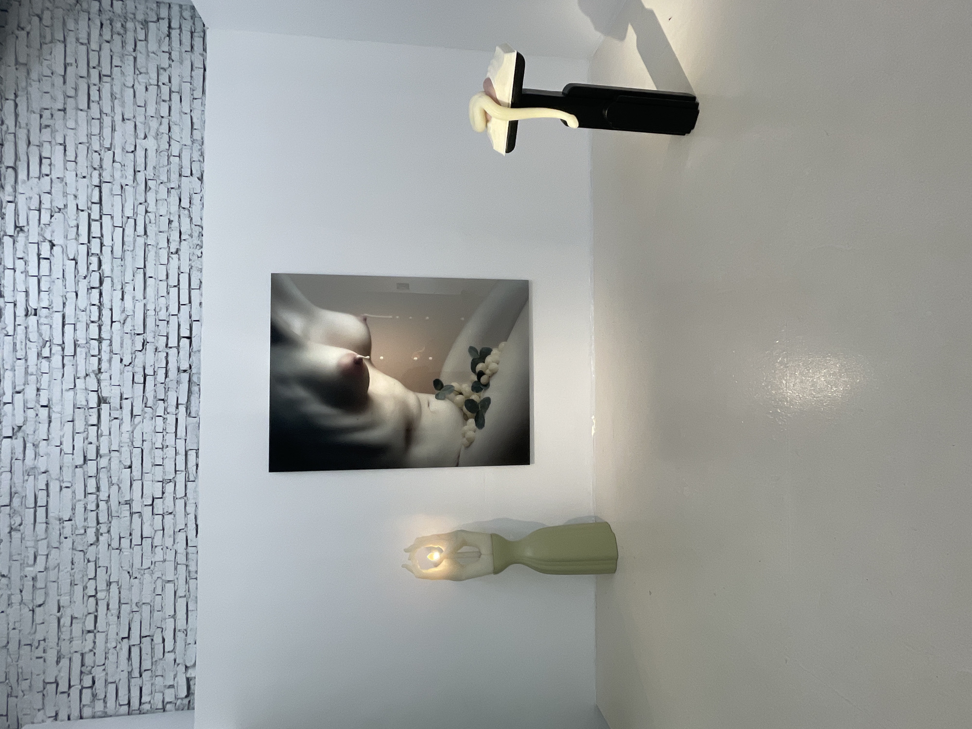 The image depicts an interior scene with white walls and a textured white brick wall on the left. Mounted on the white wall are two artistic light fixtures. The upper fixture is a black rectangular base with a pale, abstract sculptural element protruding from it. Below this, a framed artwork is displayed, featuring a close-up, soft-focus image of a person's torso with beads and leaves draped around it. Beneath the artwork, a second light fixture is visible. This fixture is pale green and has a hand-like sculpture holding a light bulb that emits a warm glow. The overall atmosphere is minimalist and artistic, with the lighting highlighting the displayed objects and artwork. The time of day and weather are not discernible from the image. No text is visible.