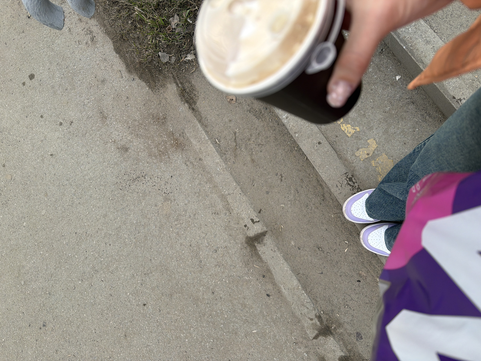 A person stands on a concrete sidewalk next to a street in Berezniki, Russia. The person is holding a cup of coffee and is wearing jeans and purple and white sneakers. A Taco Bell bag is visible. The scene appears to be taken during the day with overcast skies.