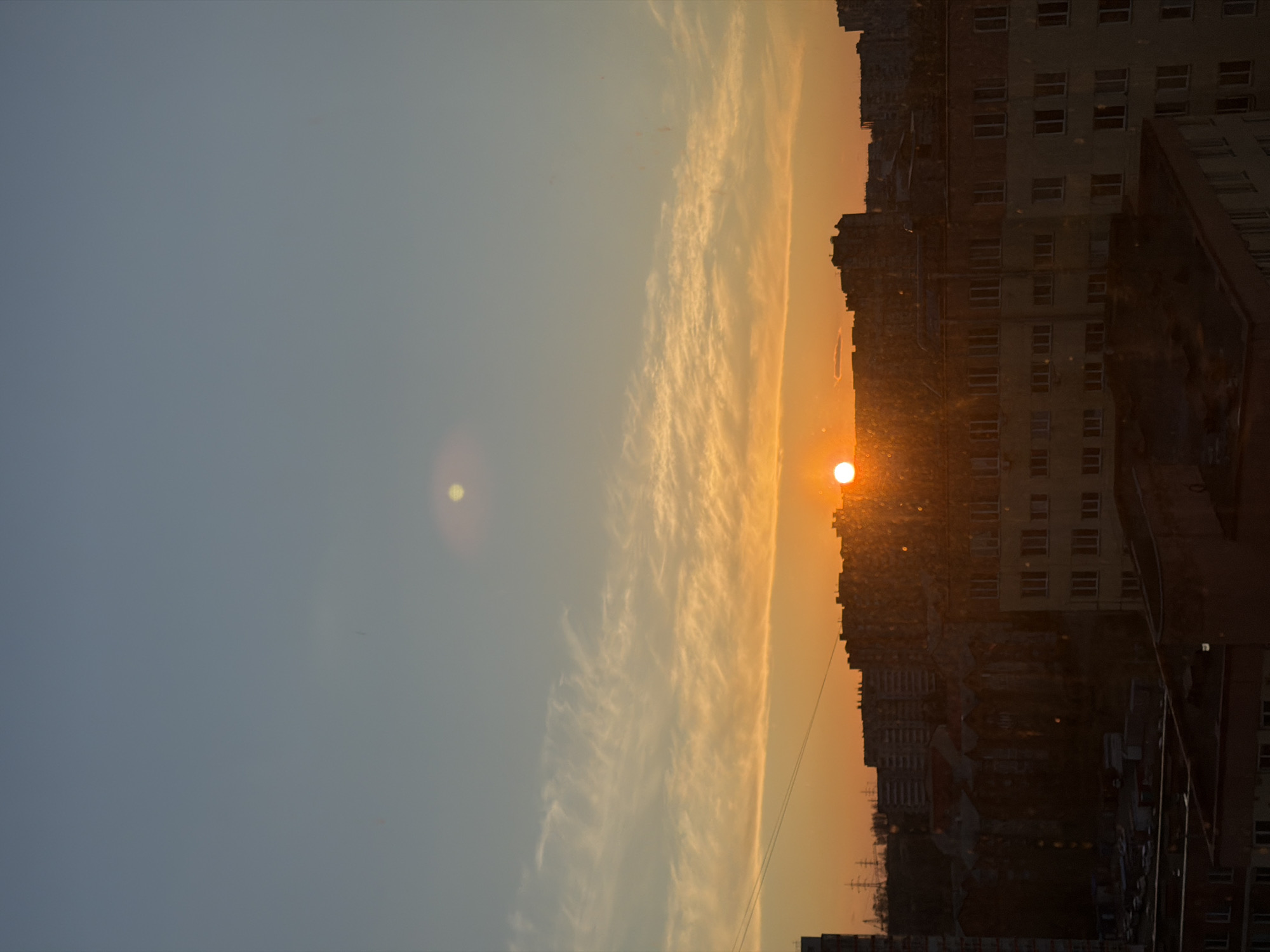 The image depicts a sunset over an urban landscape. The sky transitions from a blue-grey hue on the left to a vibrant orange as it nears the horizon on the right. The sun appears as a bright, orange orb, partially obscured by the silhouettes of buildings. The silhouettes are of various heights and architectural styles, suggesting a densely populated area. The presence of multiple apartment buildings confirms this. The city is Krasnoyarsk, Russia.