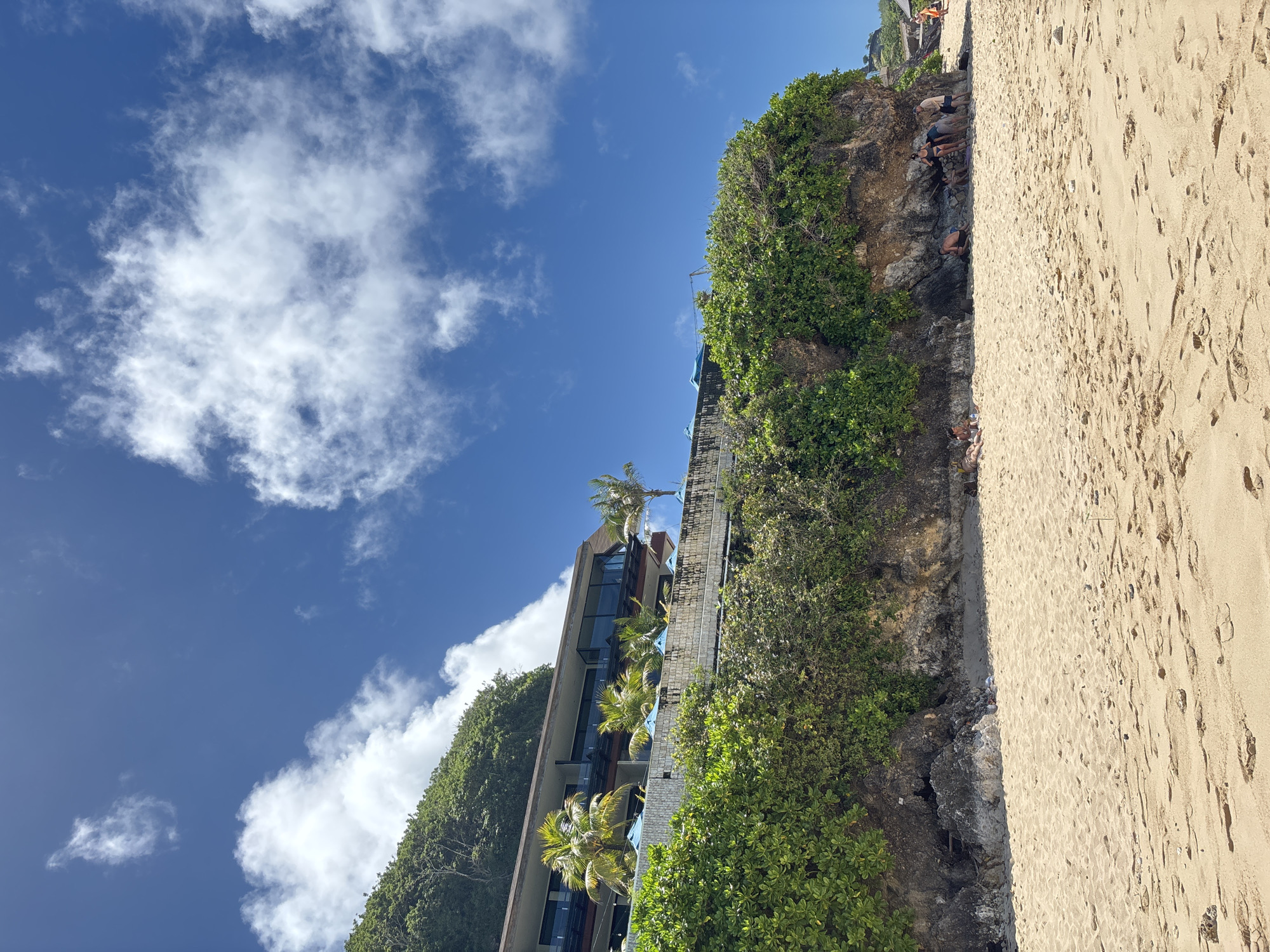 The scene is set on a beach in Ungasan, Indonesia. Sandy beach stretches to the right with footprints scattered. Adjacent to the beach is a rocky cliff covered in dense, green vegetation. Along the cliff, several people are gathered. To the left of the cliff, a modern building is visible, possibly a hotel or resort. Palm trees are interspersed within the building's structure. The sky overhead is a clear blue, dotted with white, fluffy clouds.