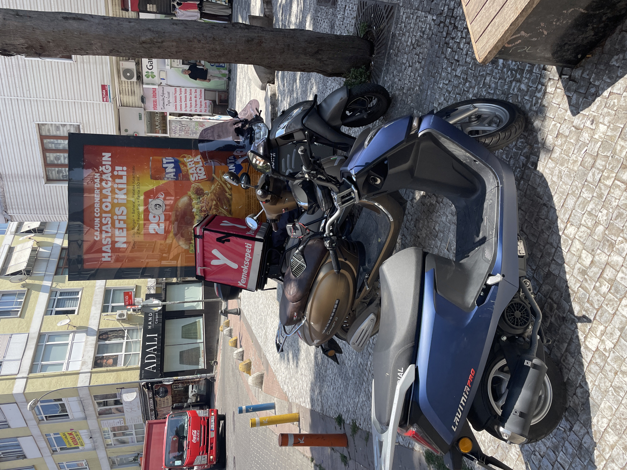 The image depicts a street scene in Kadıköy, Turkey, likely on a sunny day, with several parked scooters and a red delivery truck. In the foreground, a blue scooter with the text "LAVINIA PRO" is parked on a cobblestone sidewalk. Behind it, a brown scooter is parked, and further back, a dark gray scooter with a red delivery bag on its rear rack is visible. The delivery bag has a large "Y" logo, indicating it belongs to Yemeksepeti, a popular Turkish food delivery service.
To the left of the scooters, a red Coca-Cola branded delivery truck is parked. Behind the truck and scooters, there are buildings lining the street. One building on the left has advertisements on its facade, including one with text in Turkish and an image of a woman. A sign above a shop reads "ADALI HAND Carpet".
Further down the street, a large billboard promotes "CAJUN CORNER'S" with images of food and drinks. To the right of the billboard, a tree stands on the sidewalk, and in the background, a person is standing near a storefront. The overall scene is characteristic of a bustling urban environment.