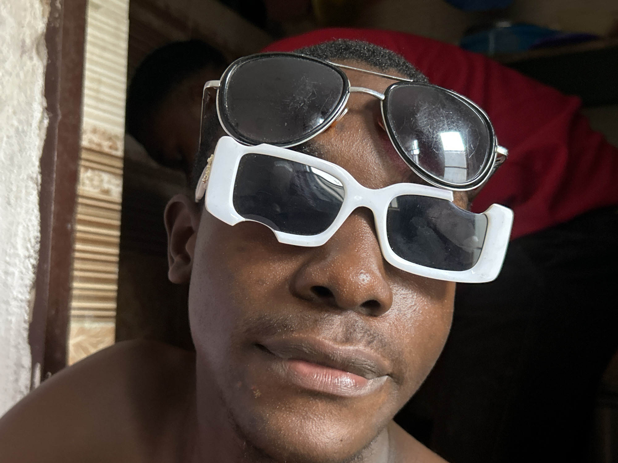 A close-up, eye-level medium shot shows a Black male wearing two pairs of sunglasses, one atop the other. The top pair is a silver-rimmed aviator style, while the bottom pair is a white, rectangular, chunky-framed style. The man is looking directly at the camera with a neutral expression. His skin appears oily, and he has a light mustache and stubble. He is shirtless, and his shoulders are visible. The background is out of focus, but it appears to be indoors, with a textured wall or screen behind him and a red garment to the right. The lighting suggests it is daytime. Based on its prevalence in Ihiagwa, Nigeria, the scene is likely a residential setting.