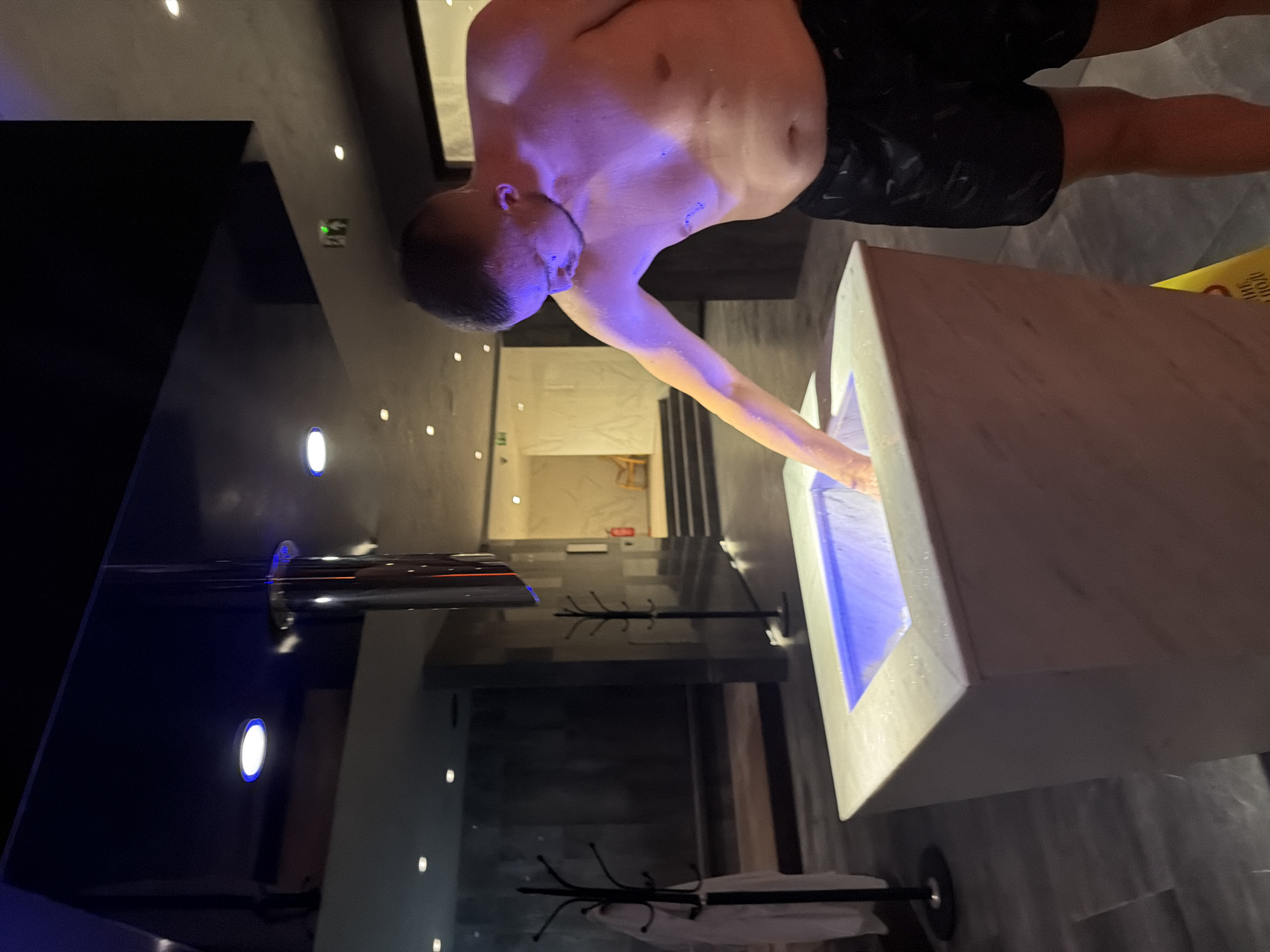 In Hisarya, Bulgaria, a shirtless man with short hair and visible water droplets on his skin is leaning over a white marble basin filled with blue-lit water. He is reaching his left hand into the basin, appearing to test the water temperature. The man is wearing black patterned swim shorts. The environment suggests a spa or wellness facility, with dark tiled flooring, modern lighting including blue accents on the ceiling, and a large reflective metallic cylinder suspended from above. In the background, there are coat racks with white robes, dark walls, and stairs leading to a brighter area with a wooden chair and a fire extinguisher. The lighting casts a purple hue on the man's upper body.