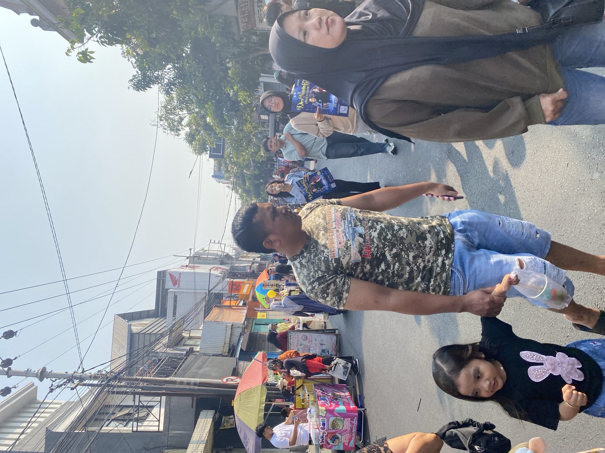 This is a street scene in Kediri City, Indonesia. A man in a camouflage t-shirt and ripped blue jeans walks forward, holding a plastic bag. Beside him, a young girl in a black t-shirt with a pink bunny design and a blue skirt holds his hand. She also holds a small plastic bag with items inside. Behind them, other people are walking on the street. Some are holding posters. The street is lined with buildings and trees. There are also several colorful umbrellas and vendor stalls visible in the background, suggesting a market or public event. The sky is clear and blue.