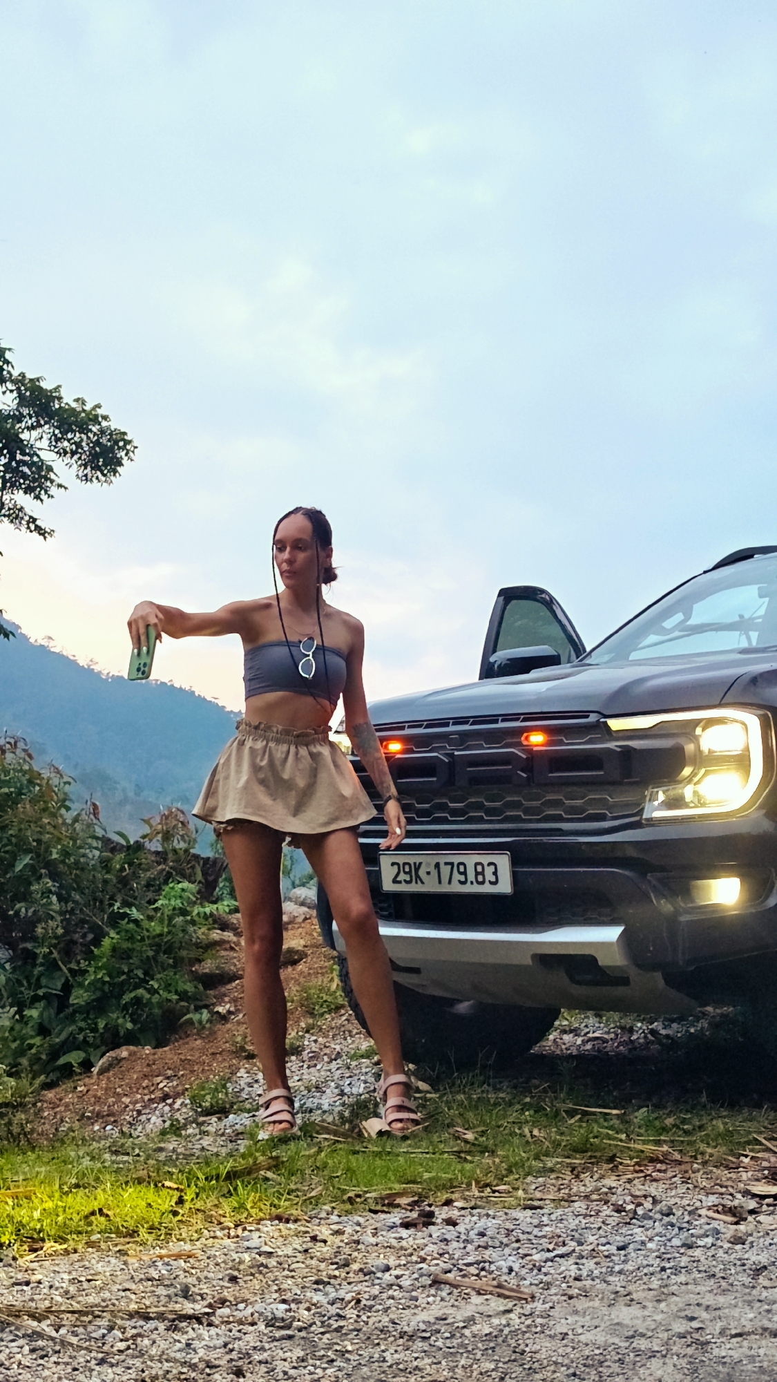 A full-body vertical shot captures a young woman standing on a gravel road beside a black pickup truck, holding a green smartphone outstretched, seemingly taking a selfie or video. She wears a grey strapless crop top, a light brown ruffled mini-skirt, and light pink strappy sandals. Sunglasses are tucked into her top, and she has visible tattoos on her left arm. Her braided hair is styled in several thin strands down her front.
The black pickup truck, identifiable by the "FORD" lettering on its grille and prominent headlights, has its headlights on, emitting a warm glow. Red lights are visible on the grille above the license plate. The Vietnamese license plate reads "29K-179.83". The driver's side door of the truck is open.
The scene is set in a mountainous, rural environment, consistent with Nậm Dịch Commune, Vietnam. The ground is a mix of gravel and dirt, with patches of green grass and bushes on the left and in the mid-ground. Distant green mountains rise in the background under an overcast or twilight sky, suggesting late afternoon or early evening. The soft, diffused light creates a serene outdoor atmosphere, hinting at a stop during a journey or an adventure.