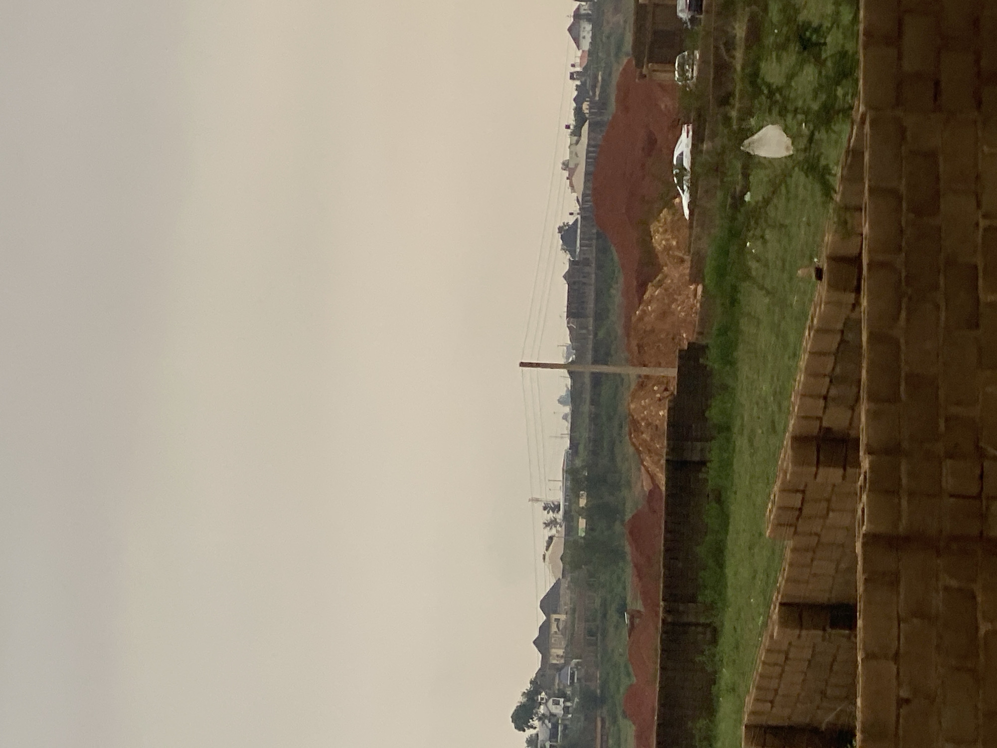 The image depicts a partially constructed brick building in the foreground, with walls of light brown bricks laid out in a row. Beyond this, a low, dark brick wall separates it from a grassy area dotted with a few small trees. In the middle ground, large mounds of reddish-brown soil are visible, suggesting ongoing construction or landscaping. Further back, a line of houses with various roof designs, some with dark, pitched roofs and others with lighter, flatter styles, are situated on a gently sloping hill. Power lines are strung across the scene between utility poles. A white car is parked near the mounds of soil, and another vehicle, possibly an SUV, is parked further to the right. The background is dominated by a hazy, overcast sky. The overall scene suggests a developing residential area in the city of Jos, Nigeria, characterized by a mix of construction and established housing.