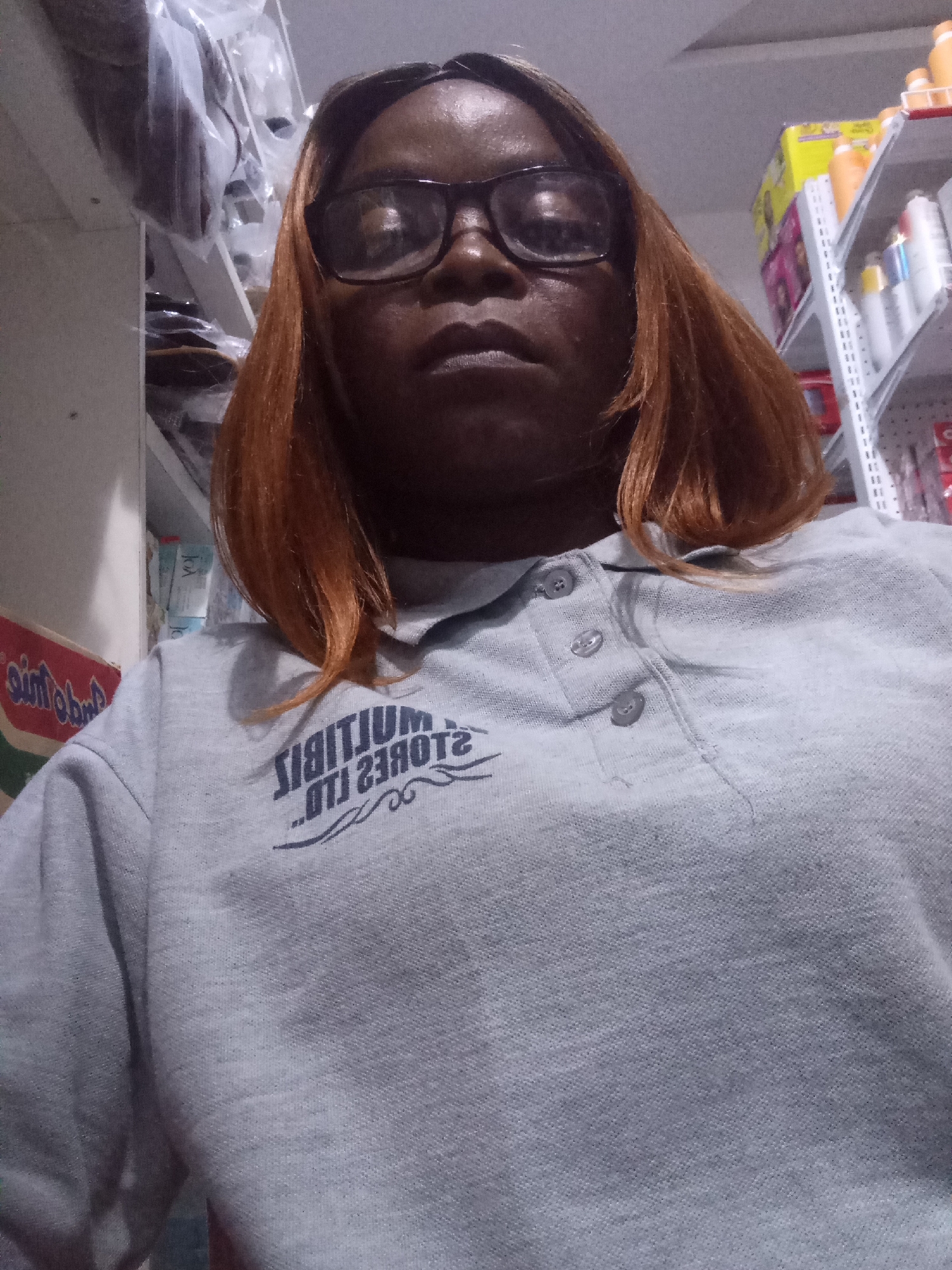 The image is a close-up, low-angle selfie of a dark-skinned woman wearing glasses and a light grey polo shirt. The woman has reddish-brown hair. The polo shirt has a logo printed on the left chest area which appears to be a reversed text that reads ".OTI 2380T2 .SIITIJUM". Behind the woman, shelves are visible, stocked with various products. On the far left, there is a portion of a red and white packaged item. On the right side of the image, shelves are filled with bottles, possibly toiletries or cleaning supplies. The overall environment suggests an indoor retail setting.