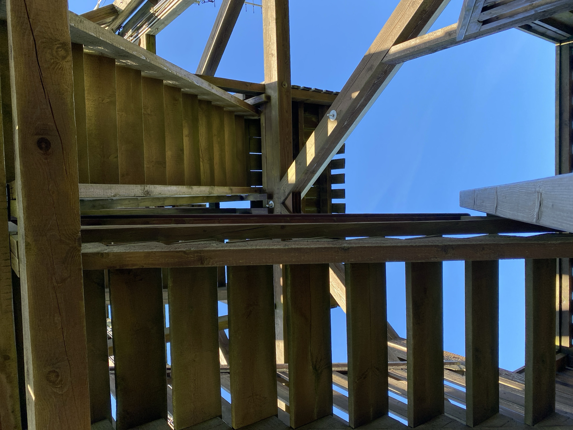 An outdoor, upward-angle shot shows a wooden staircase against a bright blue sky. The staircase is constructed from roughly hewn timbers, with visible grain and some signs of weathering. Multiple steps are visible, with gaps between them allowing the sky to show through. The structure appears to be part of a larger wooden construction, possibly a viewing tower or observation deck, with diagonal supports and horizontal beams also visible. The lighting suggests a sunny day, with shadows cast by the wooden elements. The image does not depict any people, specific landmarks of Busenhausen, Germany, or any discernible actions or interactions.