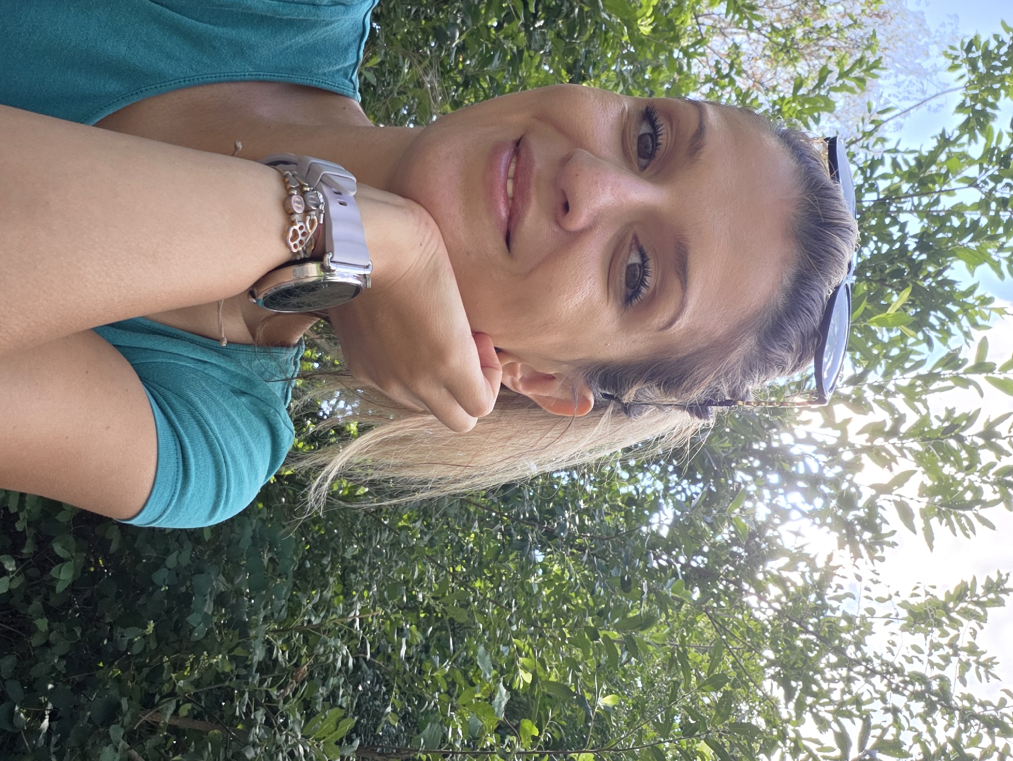 A medium shot photograph captures a woman posing outdoors, leaning her chin on her hand. She has blonde hair and is wearing a teal t-shirt and a watch with a light purple band and a silver watch face. She is also wearing a delicate bracelet with a chain-link design. Sunglasses are perched on top of her head. The background is filled with lush green trees and a bright sky, suggesting a park or a natural setting within the city of Munich, Germany. The lighting indicates it is daytime, with sunlight filtering through the foliage.