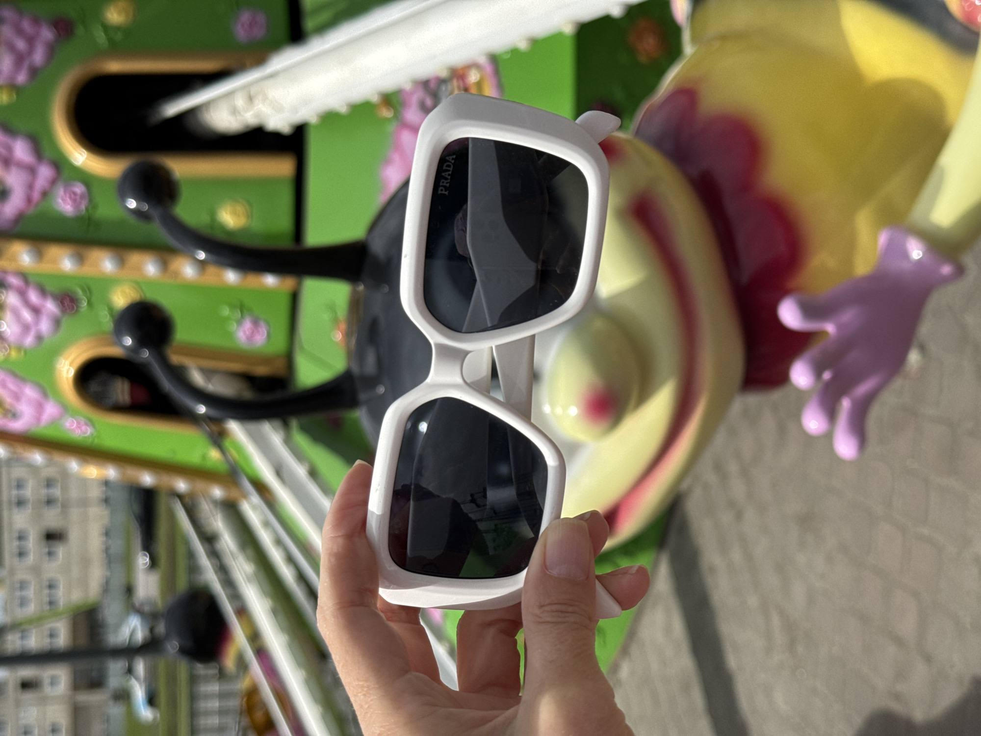 A hand holds a pair of white Prada sunglasses in front of a blurry merry-go-round figure, likely at a carnival or amusement park. The background shows a building indicating an urban setting in Saint Petersburg, Russia. The carousel figure behind the hand depicts a bee or insect-like creature with yellow coloring and a pink frill. The carousel mechanism is also partially visible with green panels and black protrusions. The sunglasses are angled to display the Prada logo on the lens. The hand is fair-skinned with manicured nails.