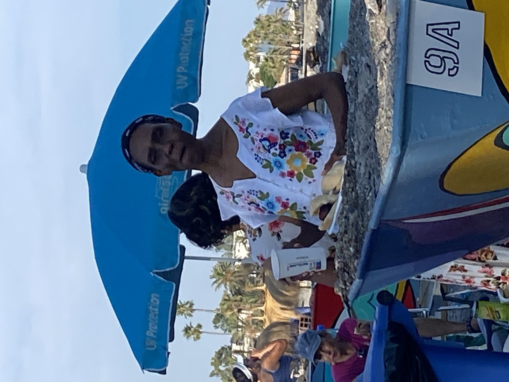 The image depicts a sunny day at a beach in Marbella, Spain. The primary subject is a person wearing a white shirt with colorful floral embroidery and a black headband. They are holding a white cup and appear to be interacting with someone or something off-camera. In the background, a blue umbrella with "UV Protection" written on it is visible. Further back, palm trees suggest a tropical or Mediterranean environment. There's also a sign with "9A" on it attached to a blue object. Other people are present in the scene, some seated and others partially visible, suggesting a gathering or a busy beach day. The overall atmosphere seems relaxed and casual, indicative of a leisure activity at the beach. The lighting suggests it is daytime.