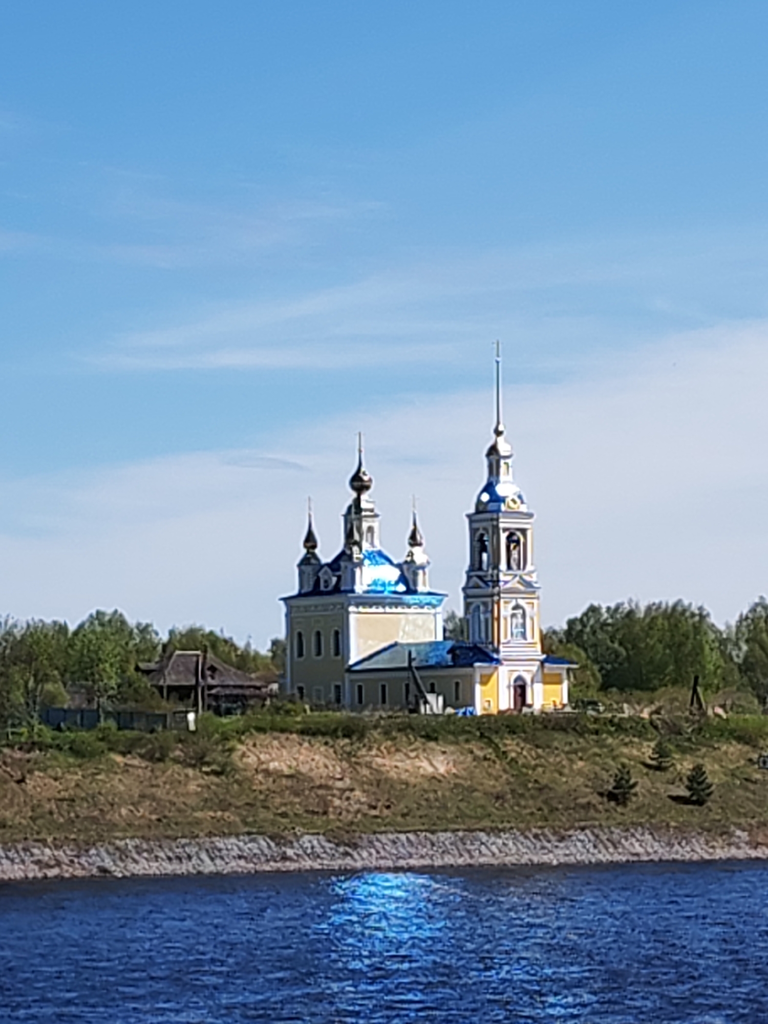 A landscape depicts a yellow Orthodox church with blue accents situated on a grassy hill overlooking a river, likely in Глебово, Russia. The church has a central building with multiple domes and a separate bell tower. The river in the foreground is dark blue with a shimmering reflection of the sun. Behind the church, there are trees and a small, wooden building. The sky is light blue with scattered clouds.