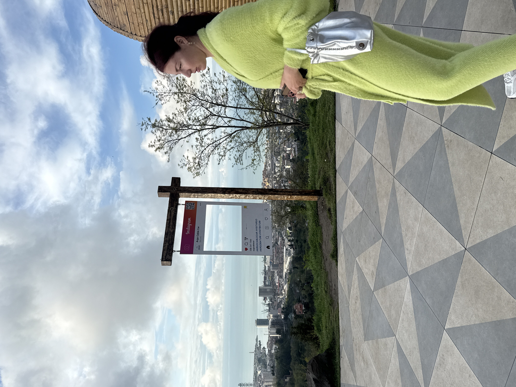 Here is a detailed description of the image:
**Scene:**
The image shows a woman standing on a geometrically patterned paved area, with a panoramic view of the city of Batumi, Georgia in the background. The weather is sunny with some scattered clouds.
**People:**
* A woman with her hair tied back is visible. She is wearing a lime green cardigan and white sneakers. A small, silver shoulder bag hangs on her left shoulder.
**Objects:**
* A wooden sign is positioned in the foreground, displaying an Instagram frame for Argo Cable Car (Argo Cable Car (Argo Batumi)). It features details such as an Instagram logo, Argo Cable Car name and profile, number of likes (598,541), account name (argocablecar) and relevant hashtags.
* The city of Batumi is visible in the distance, with buildings of various heights. The Black Sea stretches out towards the horizon.
**Environment:**
* The foreground is dominated by a grey and white geometrically patterned pavement.
* There is some greenery separating the pavement from the city below.
* The sky is bright blue with scattered clouds.
* A partially visible bamboo structure appears on the left top of the image.
**Location:**
Based on the Instagram sign for Argo Cable Car and the cityscape visible, the location appears to be at or near the Argo Entertainment Center in Batumi, Georgia, which offers a scenic viewpoint of the city and the sea.