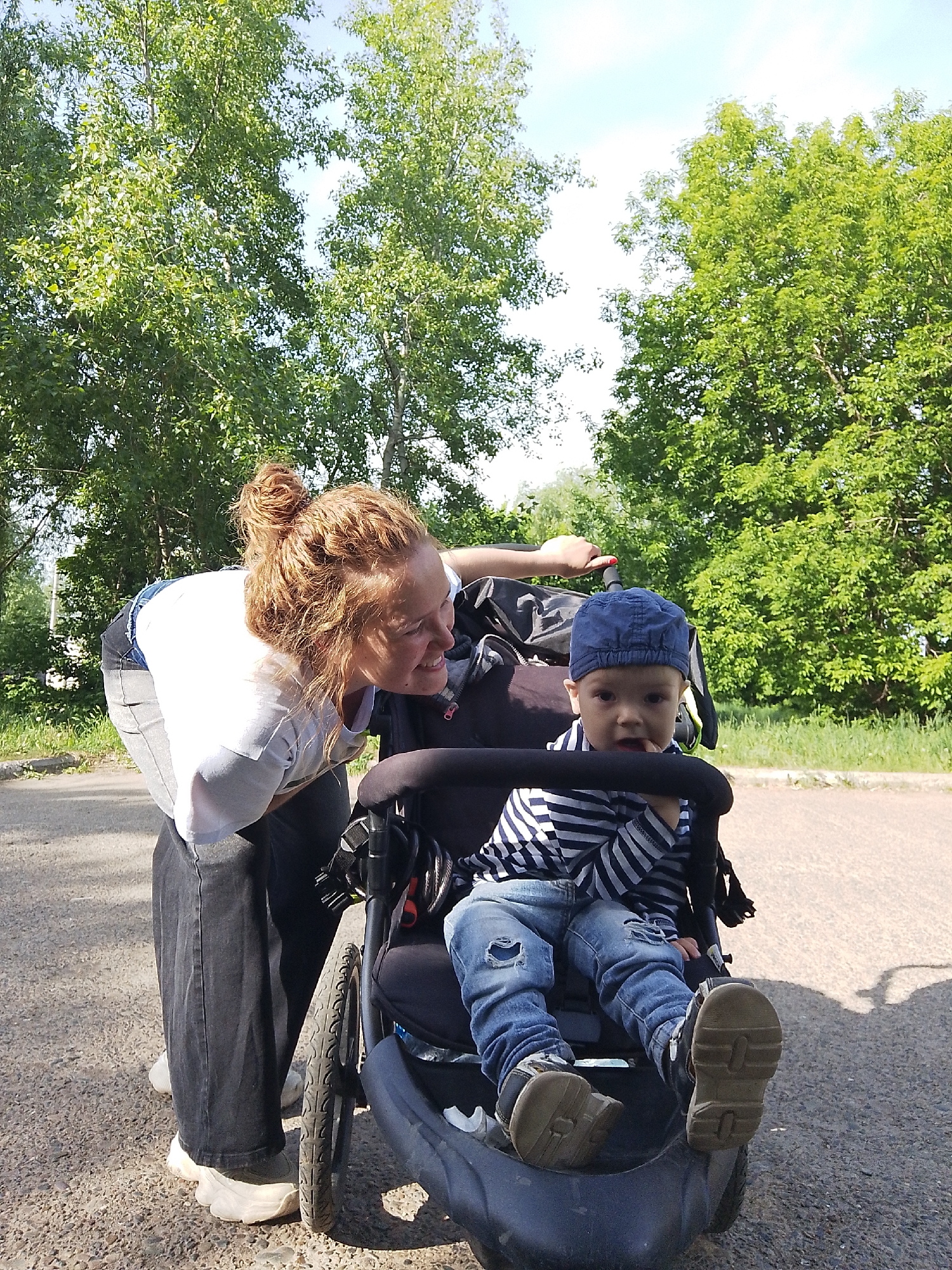 The image depicts an adult female interacting with a young child seated in a stroller on a paved surface, likely a road or walkway. The female is bending over and looking at the child, indicating an interaction. The child is wearing a cap, a striped shirt, and jeans, and is seated with their legs extended. The setting includes greenery and trees in the background, suggesting a park or outdoor area. The location is unidentified.