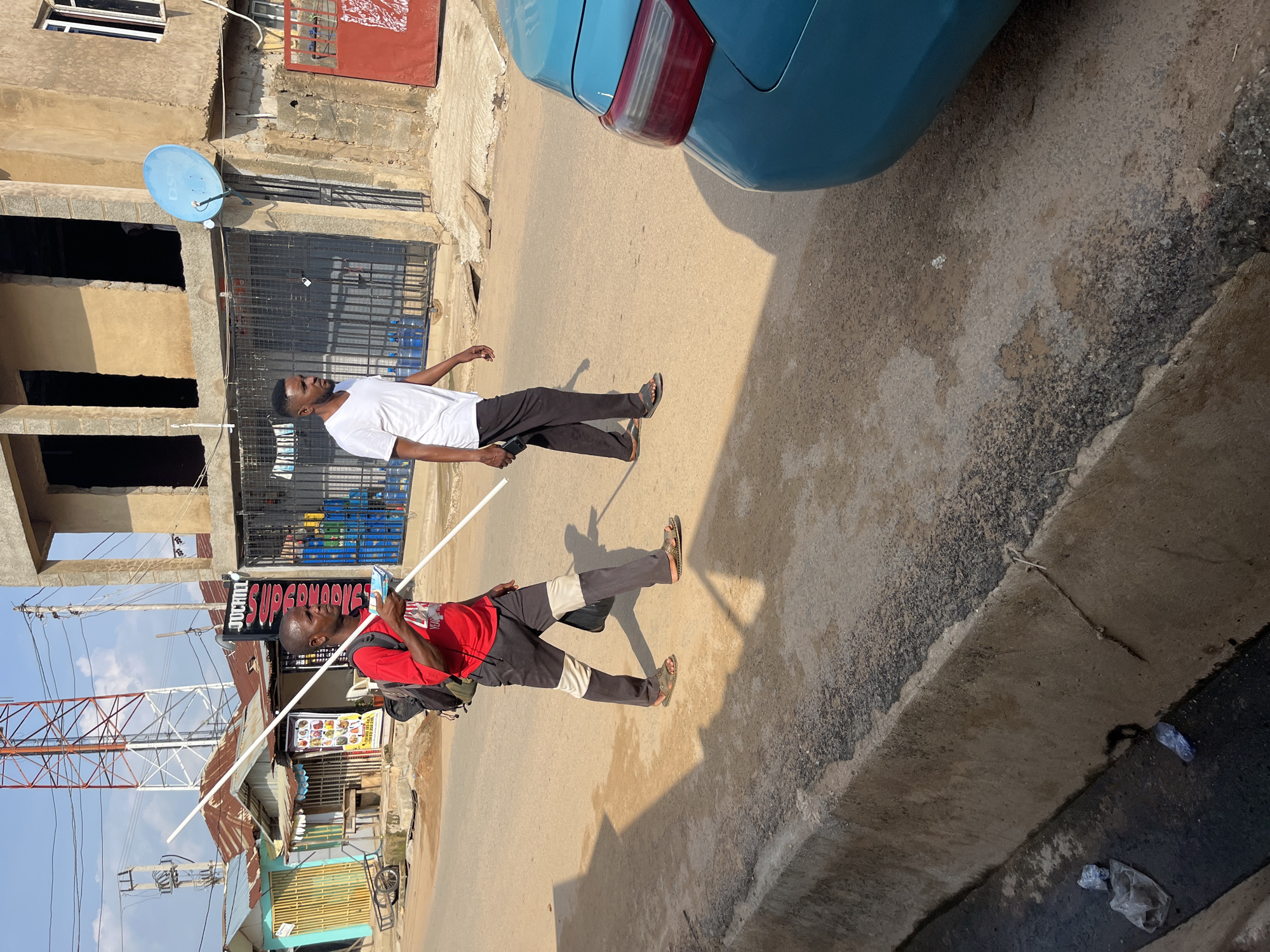 Two men walk on a paved street in Abuja, Nigeria, during daylight. The man on the left wears a red t-shirt, dark pants with cream and gray accents, and sandals. He carries a backpack and holds a long, white pipe horizontally in front of him with both hands, parallel to the ground. The man on the right wears a white t-shirt, black pants, and sandals. He walks with his hands at his sides. The street has a concrete gutter on the left side, containing some litter. Buildings line the right side of the street. One building is under construction with exposed rebar. A blue satellite dish is mounted on one of the buildings. A turquoise car is parked on the right side of the street. The sky is clear and blue.