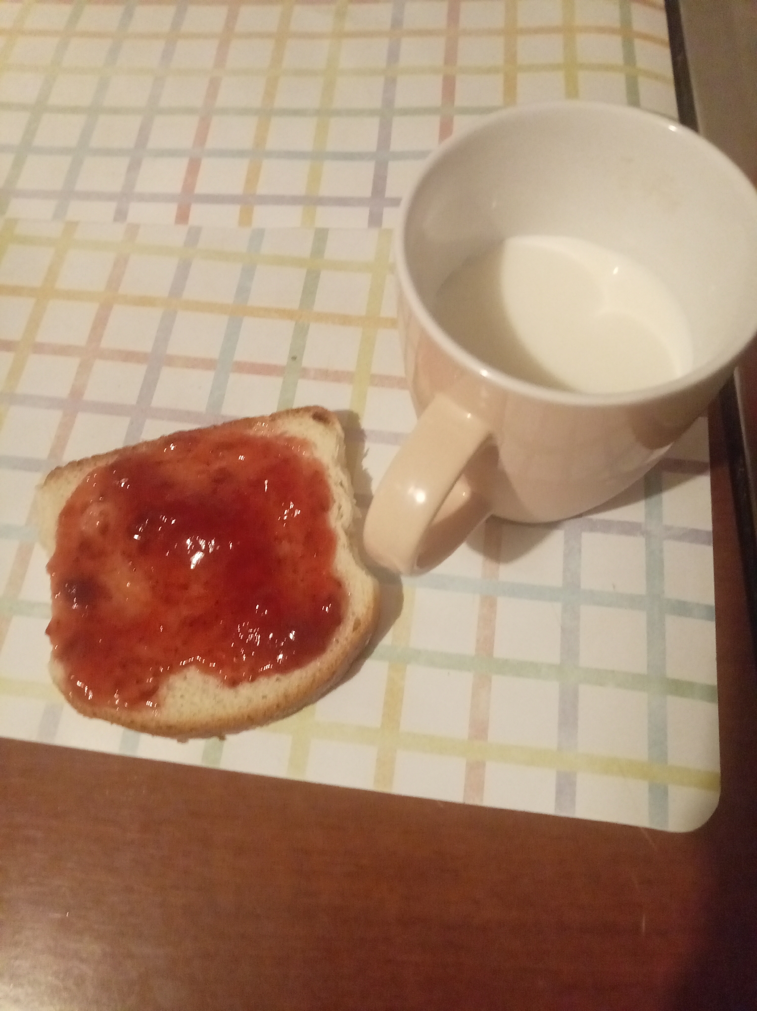 A slice of bread topped with strawberry jam sits on a placemat next to a mug of milk. The placemat has a checkered pattern of pastel colors. The bread and jam are in the lower left of the image, and the mug is in the upper right. The mug is pale pink and appears to be made of ceramic. The milk inside is white and opaque. The scene is set on a brown wooden table. The image is taken from a slightly overhead angle. The location is identified as Tapa, Estonia.