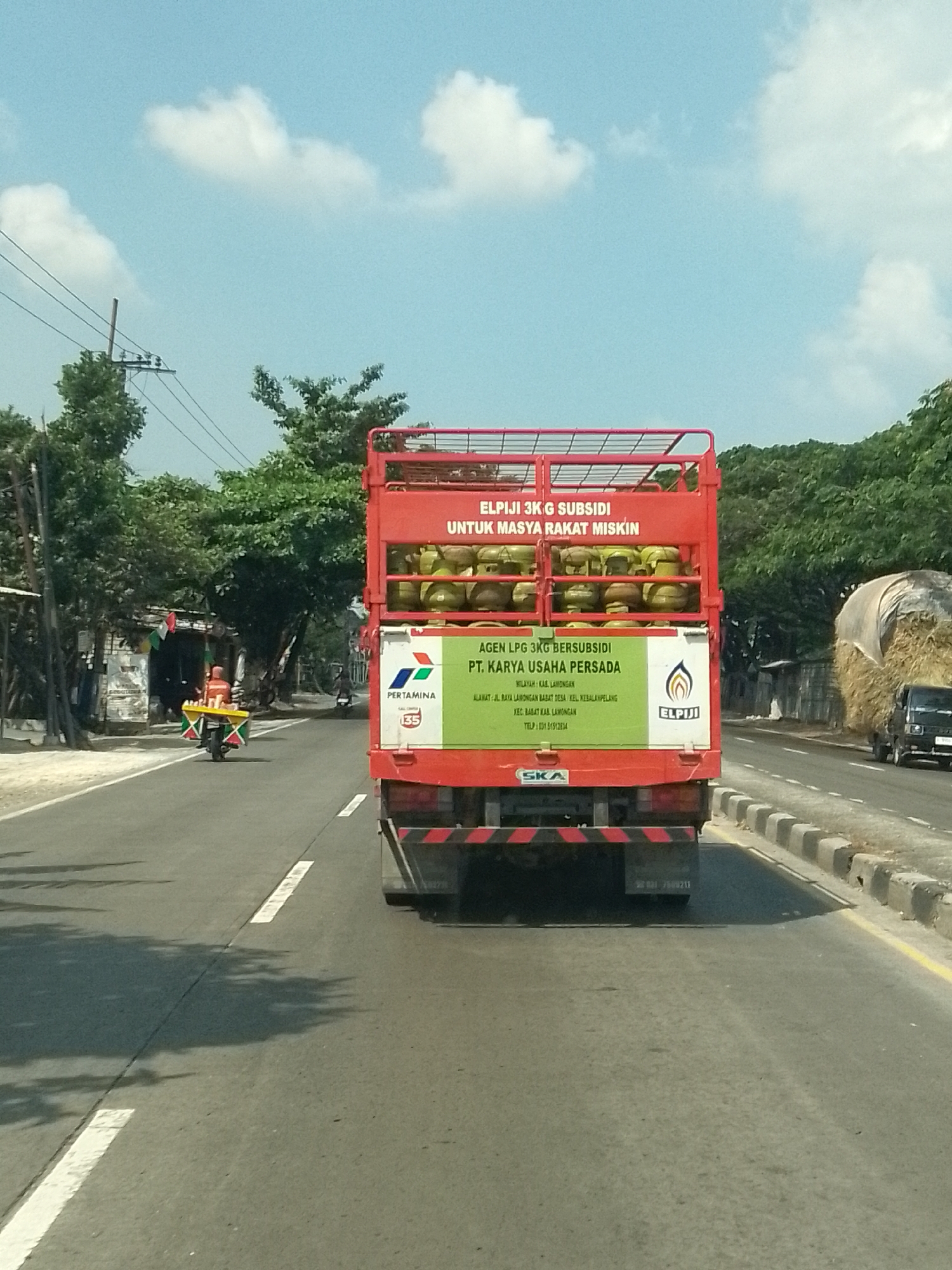 The image depicts a rear view of a red truck carrying LPG gas cylinders on a sunny day. The truck is on a road in Tritunggal, Indonesia. The side of the truck has green signage that reads "AGEN LPG 3KG SUBSIDI PT. KARYA USAHA PERSADA" and lists the agency's address and phone number. The truck is laden with numerous yellow LPG cylinders stacked inside. In the background, there are trees, power lines, and some buildings. To the left of the truck, a person is riding a motorcycle with a cart attached, also carrying LPG cylinders. Further down the road, a hay bale is visible next to a black truck. The sky is blue with scattered white clouds.