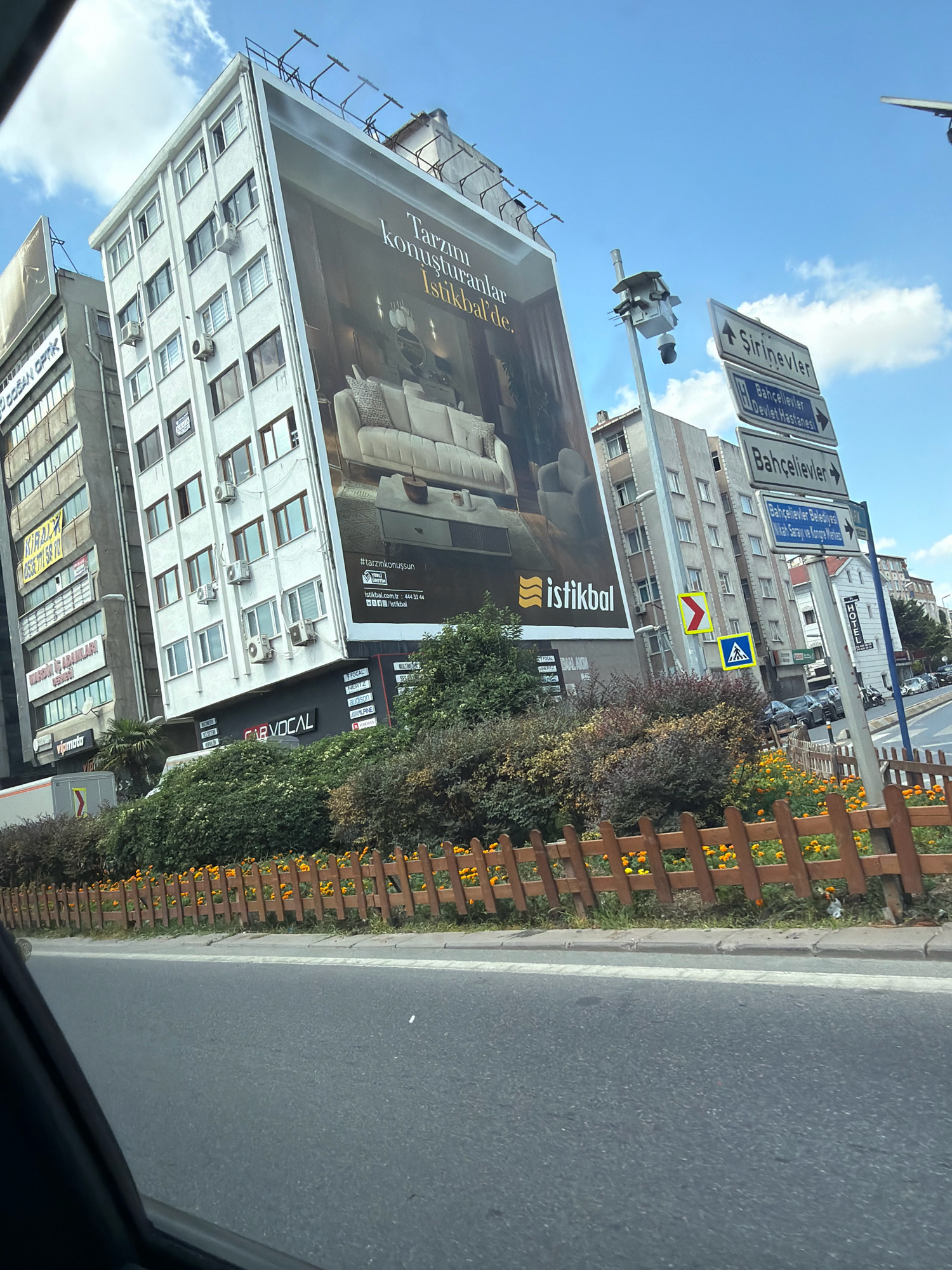 The image captures a street view in Bahçelievler, Turkey. The scene is dominated by a large building with a prominent advertisement billboard on its facade, featuring a sofa and text in Turkish. Adjacent buildings are also visible, displaying various signs and windows. A street signpost with multiple directional arrows indicating "Şirinevler," "Bahçelievler Devlet Hastanesi," and "Bahçelievler Kültür Sarayı ve Kongre Merkezi" is present. Below the billboard and on the side of a building, several smaller signs are visible, including one for "CAR VOCAL." The foreground shows a paved road with a white line marking, and a wooden fence with orange flowers is lining the roadside. The sky is blue with scattered clouds, and trees and bushes are integrated into the urban landscape. Several cars are seen parked and driving on the road in the background.