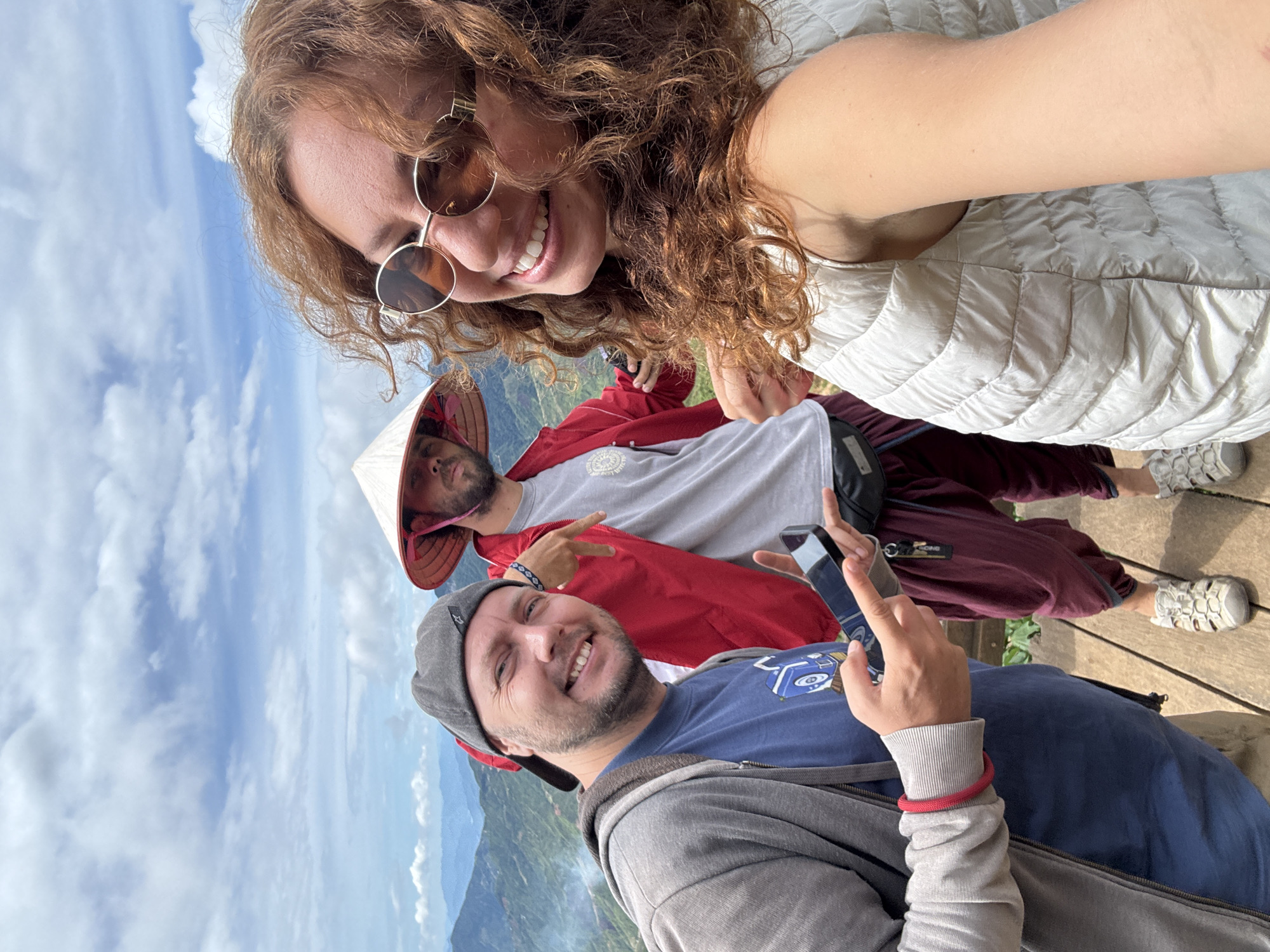 Three individuals are captured in a selfie on a wooden platform overlooking a mountainous landscape in Ta Xua, Vietnam. The person in the foreground on the right is a woman with curly, reddish-brown hair and sunglasses, wearing a white quilted vest. To her left is a man wearing a grey baseball cap, a blue t-shirt with a graphic, and a grey zip-up hoodie. He is smiling and holding a smartphone in his left hand. Behind them, a man wearing a conical hat and a red jacket is posing with his right hand pointing downwards. The background features rolling green hills under a cloudy sky, indicative of the mountainous terrain of Ta Xua, known for its breathtaking views and "cloud hunting" tourism. The platform appears to be made of wooden planks.