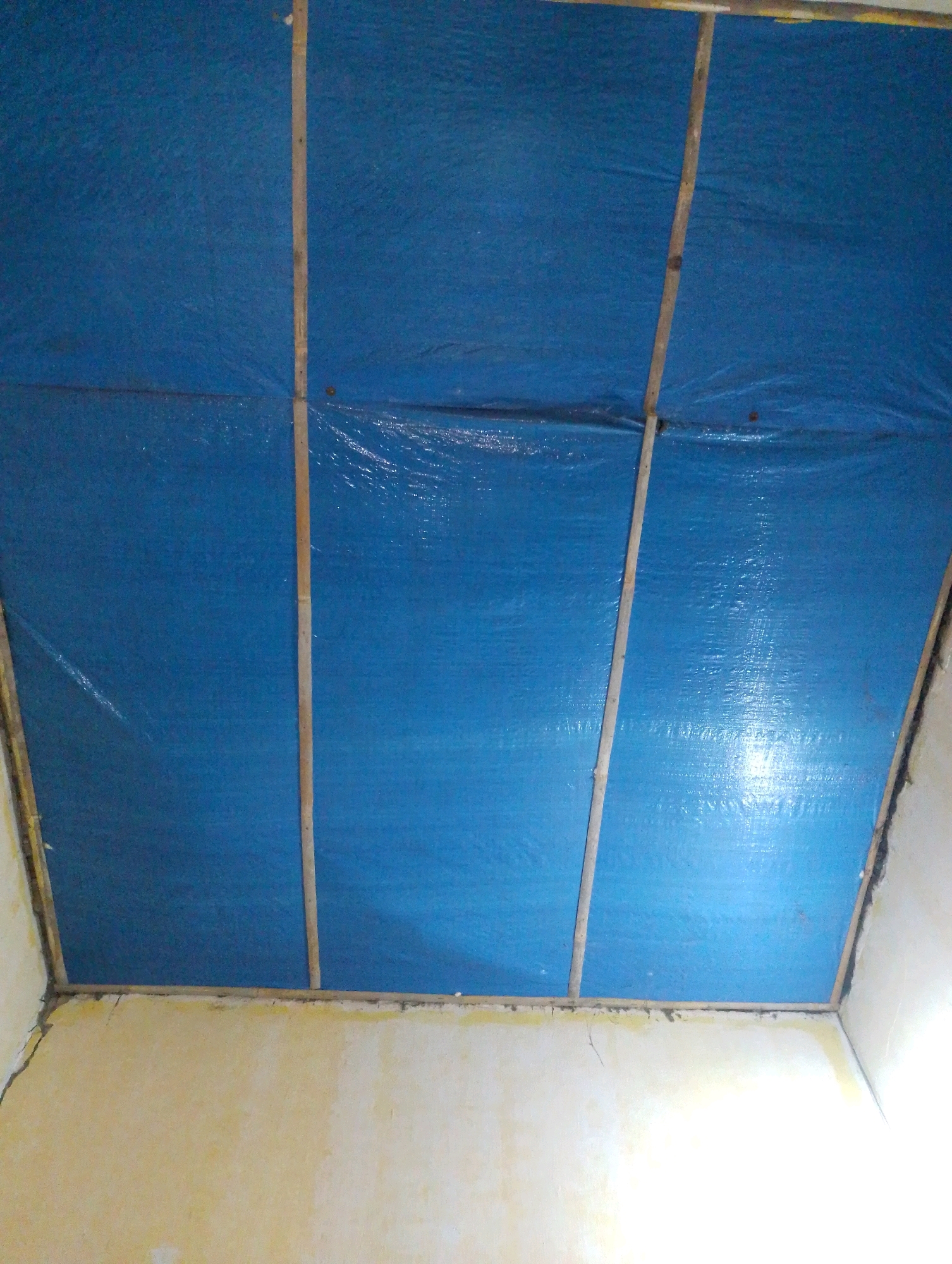A blue tarp is stretched across a ceiling, secured by thin wooden slats. The tarp appears to be in panels, with a grid-like structure formed by the slats. The walls below are a light yellow color and show signs of wear and possibly some cracking. The lighting in the scene is somewhat dim and uneven, with some areas brighter than others, suggesting interior lighting. The overall impression is that of a makeshift or temporary ceiling repair or covering. The image was captured in Plosoklaten, Indonesia.