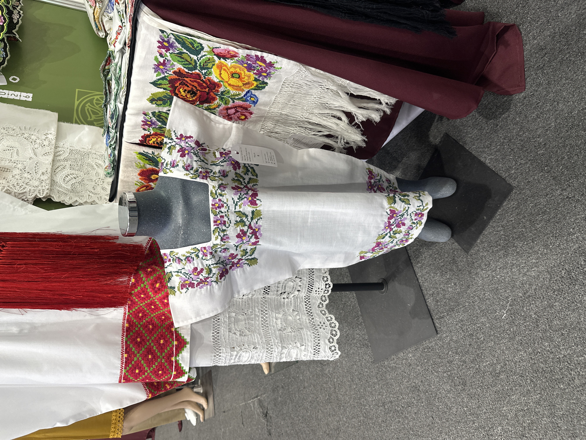 The image depicts a display of embroidered textiles, possibly clothing items, in a setting that could be a market or exhibition. The foreground features a dress draped over a mannequin, with intricate floral embroidery around the neck and hem. A white fabric with red geometric embroidery and long red fringes is visible to the left of the dress. To the upper right, there is a white textile with colorful flower embroidery and fringe, draped behind the mannequin. A maroon fabric is also present in the background. The setting is indoors, with a gray carpeted floor. Based on the types of textiles and embroidery displayed, it is possible that this image was taken in Mexico City, Mexico, as these styles are often associated with Mexican traditional clothing and crafts. There are no people visible in the image.