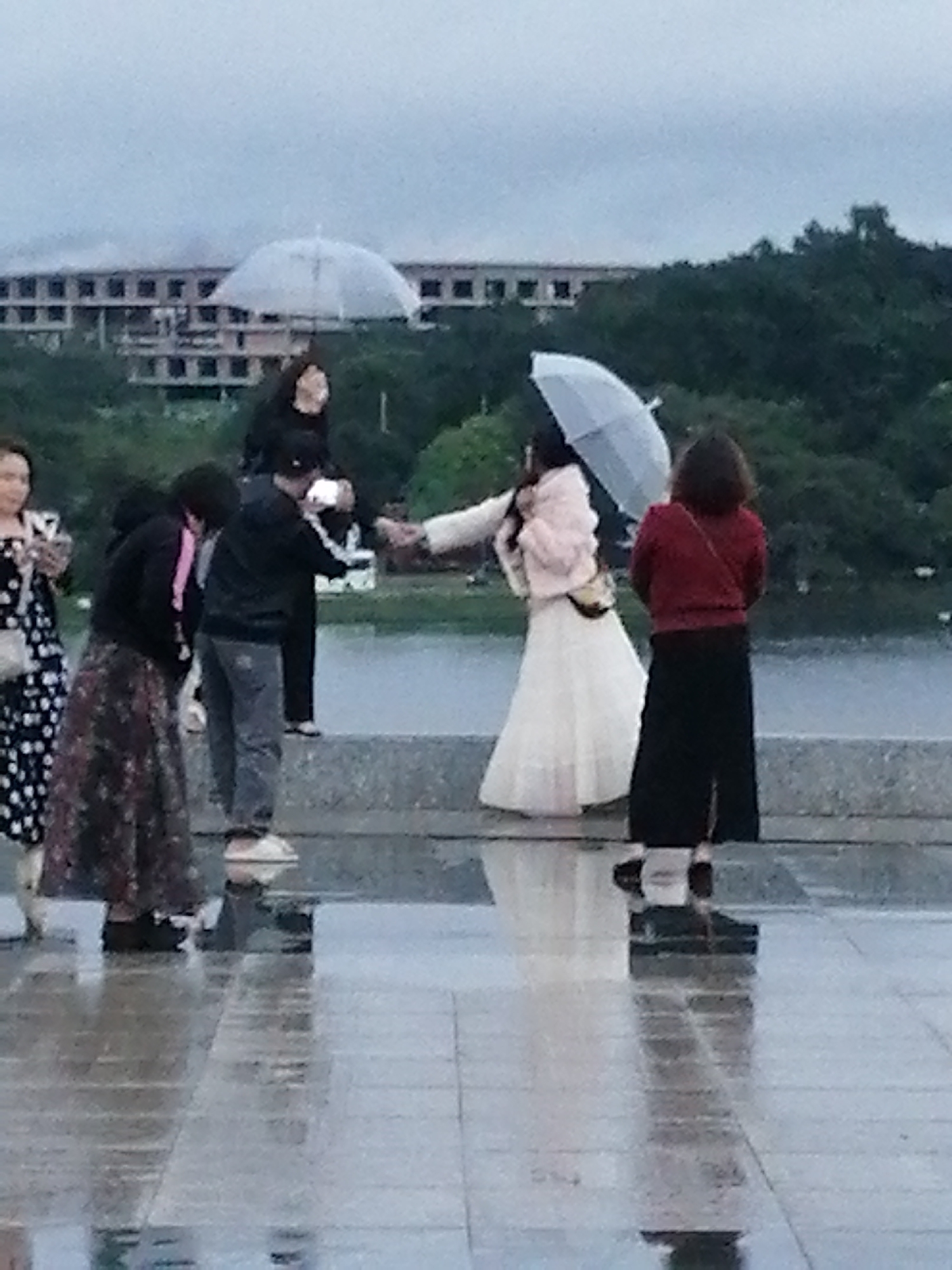 The image depicts a group of people on a wet promenade next to a body of water. In the background, there is a building complex with multiple stories and a treeline. The sky is overcast.
In the foreground, several individuals are visible. On the left, a woman in a black and white polka dot dress stands holding a bag. Next to her, a woman in a patterned skirt and dark jacket is looking down. Further to the right, two figures in dark clothing are interacting, with one holding a white object that appears to be a phone or a camera. In the center, a woman in a white dress and a light pink furry jacket is holding a light-colored umbrella. To her right, a woman in a red jacket and black pants stands facing away from the camera, with a crossbody bag.
The ground is reflective, indicating it is wet from rain. The overall scene suggests an outdoor gathering on a cloudy or rainy day. The location is identified as near Xuan Huong Lake in Da Lat, Vietnam, with the building in the background possibly being the Da Lat Railway Station or a similar architectural structure in the vicinity.