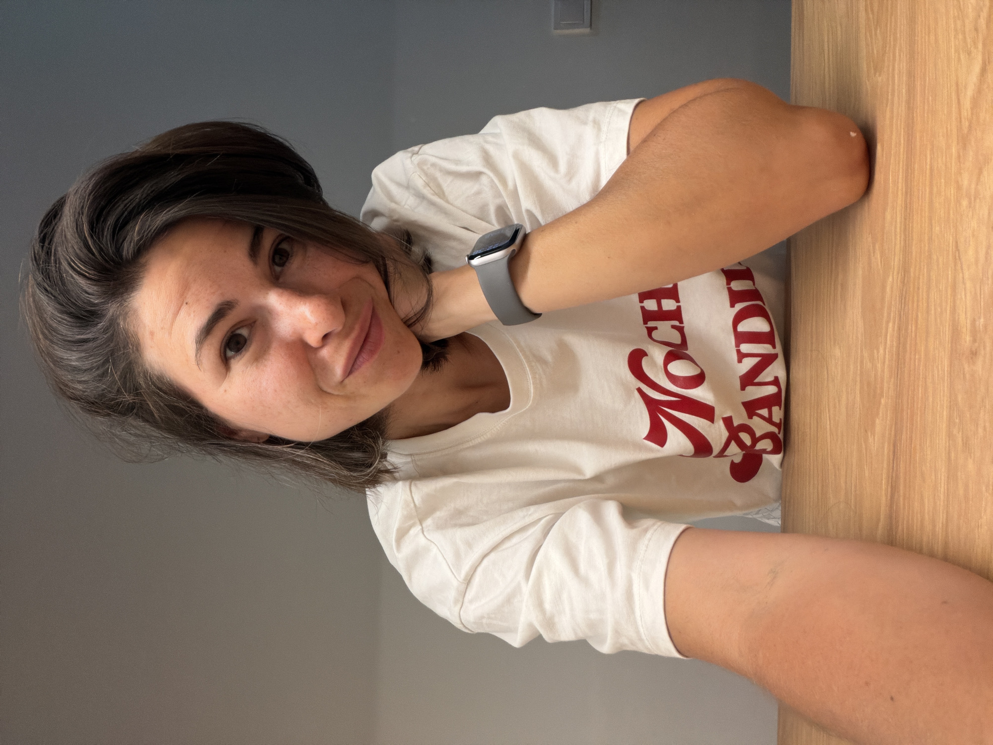 A woman poses for a selfie indoors. She has short brown hair and is wearing a white t-shirt with red lettering. A gray smartwatch is visible on her wrist. The background includes a light gray wall and a light brown wooden surface, possibly a table or countertop. The location is potentially Budva, Montenegro, but this is not verifiable from the image alone.