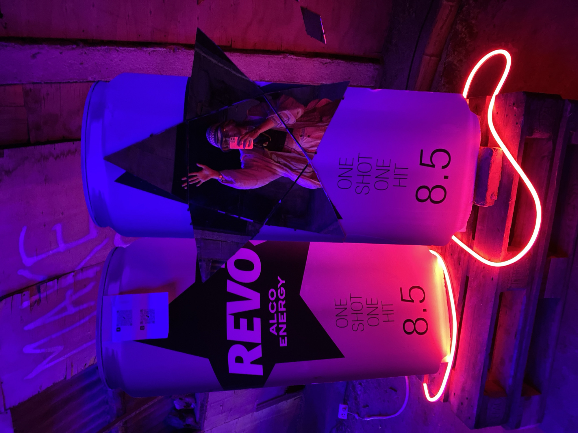 In Asureti, Georgia, a dimly lit interior space is illuminated by vibrant pink and purple lighting. Dominating the foreground are two large, cylindrical displays resembling oversized cans. The display on the left is emblazoned with the word "REVO" in large, bold purple lettering above "ALCO ENERGY" and the text "ONE SHOT ONE HIT" followed by the number "8.5". A red neon light snakes around the base of this display. The display on the right is a mirror image of the left, also featuring "ONE SHOT ONE HIT" and "8.5", but the "REVO" logo is obscured by a large, fractured star-shaped mirror. This mirror reflects a person holding a pink smartphone and waving with their left hand, wearing a patterned hat and a light-colored jacket. Both displays are mounted on wooden pallets. Above, graffiti reading "MAKE" is visible on the wooden ceiling. Electrical outlets and cords are present on the left side of the scene.