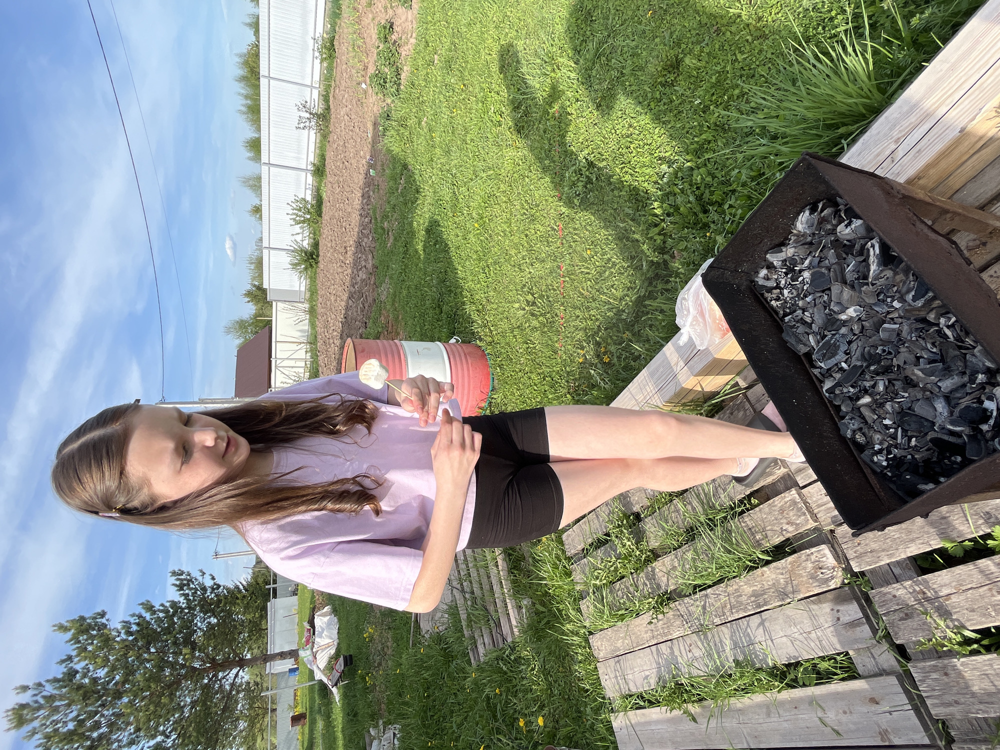 A teenage girl is sitting on wooden planks next to a barbecue grill filled with charcoal. The girl is wearing a light purple t-shirt and black shorts, and she is holding a stick with a marshmallow on it. Behind her is a grassy yard, a red and white barrel, and a white fence. A tree is visible to the left. The location is Гаревая, Russia.