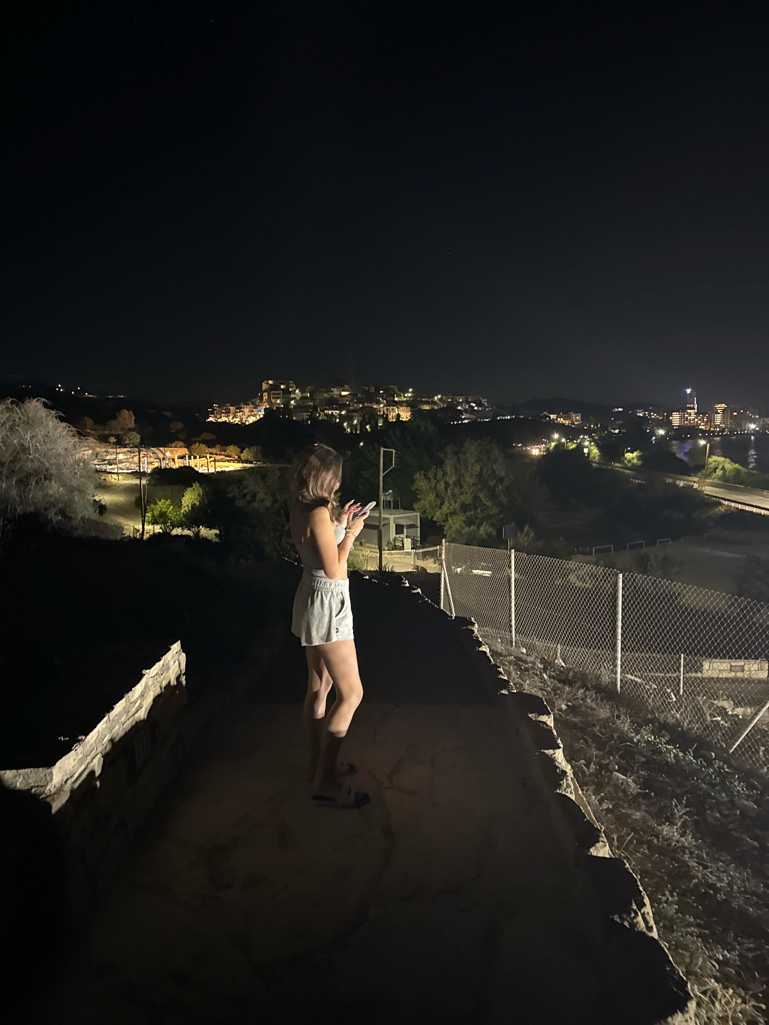 In the city of Agios Tychona Community, Cyprus, a young woman stands on a raised dirt path overlooking a town illuminated at night. She is wearing a light-colored tank top and shorts, and is holding a mobile phone in her hands. The path she stands on is bordered by a stone wall on the left, and a chain-link fence on the right. The town in the background is densely populated with buildings of various heights, many of which are lit, creating a vibrant urban scene against the dark night sky. Trees and other vegetation are visible between the path and the town, adding a natural element to the otherwise man-made landscape.