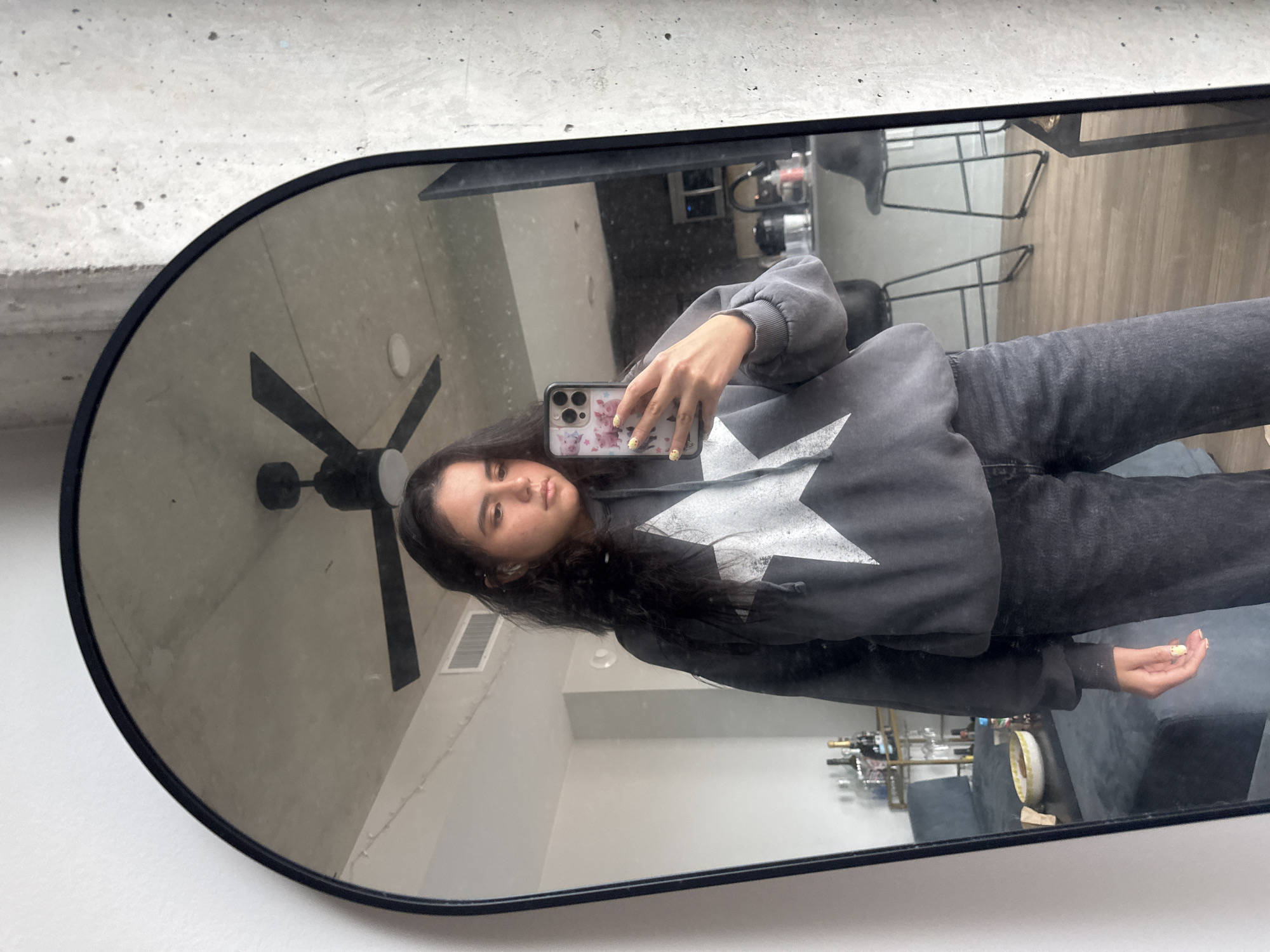 The image displays a mirror selfie taken by a young woman in what appears to be an apartment or modern living space. The woman, with long, dark hair, looks directly into her smartphone camera with a neutral expression. She is wearing a dark grey hooded sweatshirt featuring a large, distressed white star design on the front, paired with dark grey or black jeans. Her fingernails are painted a light yellow or gold, and her smartphone is in a patterned case adorned with small, light-colored cartoon animal figures, possibly pigs.
The reflection is captured in a large, rounded rectangular mirror with a black frame. The background visible in the mirror reveals interior details. To the left, a concrete wall or ceiling is visible, along with a black three-bladed ceiling fan and a rectangular ventilation register. To the right, the room features light-colored wooden flooring and a dark-colored sofa or couch. A bar cart or console is partially visible below the sofa, holding several bottles, likely wine or liquor, and various glasses. A round, light-colored decorative bowl or plate rests on the sofa or an adjacent surface. Another piece of furniture with a metal frame is seen behind the woman's arm.
The lighting suggests it is daytime. No specific weather conditions or overt emotions are discernible. The architectural elements like concrete and wood flooring point to a contemporary interior. No legible text is visible within the image. Based on the provided context, the location is Austin, United States of America.