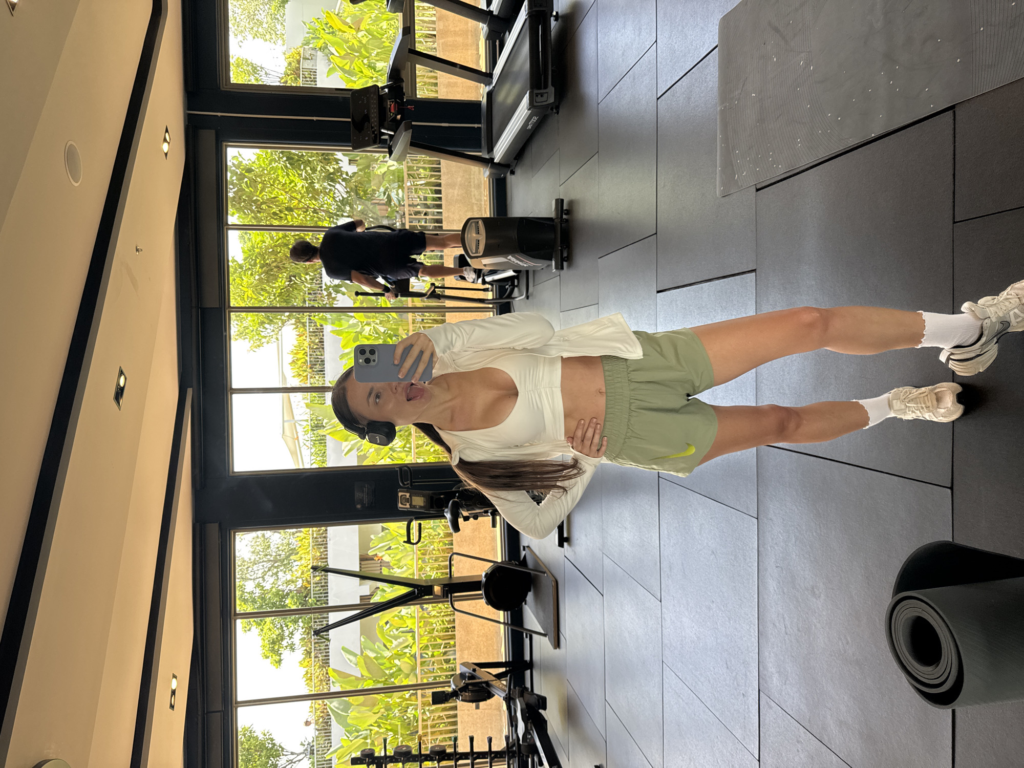 A young woman takes a selfie in a gym. She is wearing a white top, green shorts, white socks, and sneakers, and has headphones on. Her tongue is sticking out. She is standing in front of a wall of windows that look out onto a tropical landscape. Behind her, a man is exercising on a rowing machine. There are treadmills and weights visible in the gym. The gym is located in Choeng Thale, Thailand. A rolled yoga mat is on the floor.