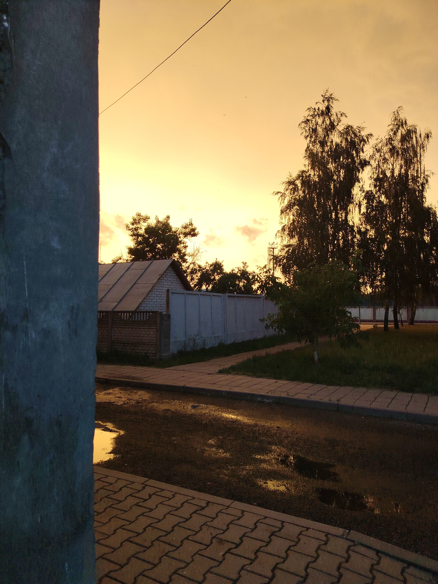 The image depicts a quiet street scene during what appears to be sunset or sunrise, casting a warm, orange glow across the sky. In the foreground, a rough, dark-colored pillar occupies the left side of the frame, partially obscuring the view. A wet asphalt road with visible puddles reflecting the sky stretches across the lower portion of the image, bordered by a patterned walkway of interlocking pavers.
Further back, a sidewalk runs parallel to the road, with a strip of green grass and foliage separating it from a light-colored fence. Behind the fence, a small, white brick building with a gray, sloped roof is visible. Several tall trees with dense foliage are silhouetted against the brightly lit sky, their branches reaching upwards. The overall impression is one of a tranquil, possibly residential, area. The specific location in the city of Brest, Belarus, cannot be determined from the visual information provided. There are no people or animals visible in the scene.