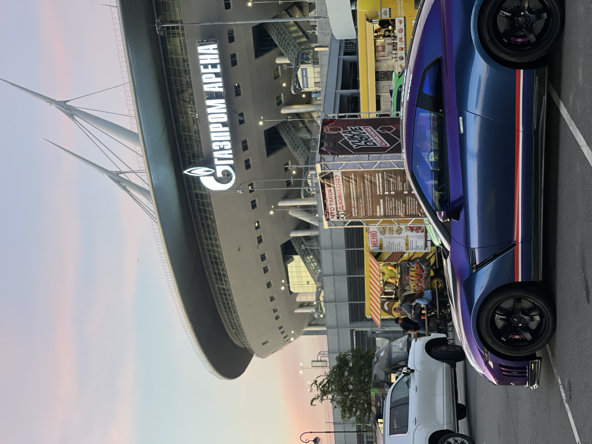 In an outdoor setting, a purple and blue sports car is parked in the foreground, angled to the right. The car has a metallic finish that shifts between shades of purple and blue, with red and white stripes along its side. The car's wheels are black, and the brake calipers are purple. Behind the sports car, a white SUV is parked to the left. Further in the background, several food trucks are set up, with people visible at some of them. One food truck has a yellow and red striped awning, and a sign with Cyrillic text. In the far background, a large, modern stadium with distinctive white spires and a large illuminated sign that reads "ГАЗПРОМ АРЕНА" (Gazprom Arena) dominates the scene. The sky above is a soft gradient of pale blue and pink, suggesting dusk or dawn. The ground is paved, and a white line marking a parking space is visible on it.