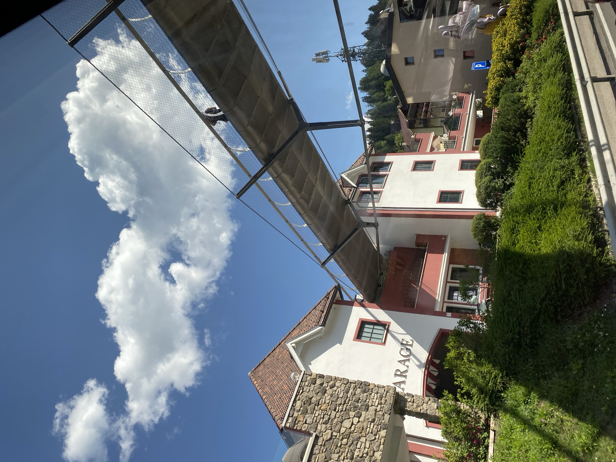 The image shows a street scene in Ortisei, Italy, on a clear day with a bright blue sky and scattered white clouds. A modern pedestrian walkway with a metal railing and mesh netting spans diagonally across the frame. A single person is visible walking on the walkway. Below the walkway are buildings with traditional alpine architecture, featuring red-tiled roofs and white or light-colored facades with red trim. One building has the word "GARAGE" prominently displayed. Green landscaping, including bushes and trees, lines the street in the foreground, with a guardrail separating it from the road. In the background, a telecommunications tower and more buildings are visible against a backdrop of a tree-covered hillside. The lighting suggests it is daytime, likely late morning or early afternoon, given the shadows.
