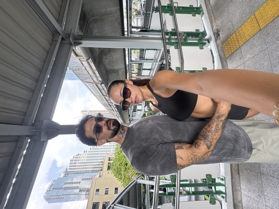 An adult male and an adult female are captured in a selfie on an elevated outdoor platform, indicative of a BTS Skytrain station or pedestrian bridge in Bangkok, Thailand. The male, positioned on the left, has dark hair, a beard, and wears dark rectangular sunglasses, a faded charcoal short-sleeved t-shirt, and light-colored shorts. Visible tattoos include a design on his neck and a full sleeve on his right arm featuring a spiderweb, a butterfly, and geometric patterns. His right arm is around the female's waist. The female, positioned slightly forward on the right, has dark hair pulled back into a ponytail, wears dark rectangular sunglasses, a black high-neck sports bra or crop top, and black shorts. Her right arm is extended, holding the camera. She looks directly at the camera, while the male looks slightly past it. The background features modern high-rise buildings under a bright, partly cloudy sky. To the right, an elevated concrete railway structure is visible. The platform itself has grey tiled flooring with yellow tactile paving on the right, bordered by metal railings with green and white panels. Grey metal structural beams are overhead.