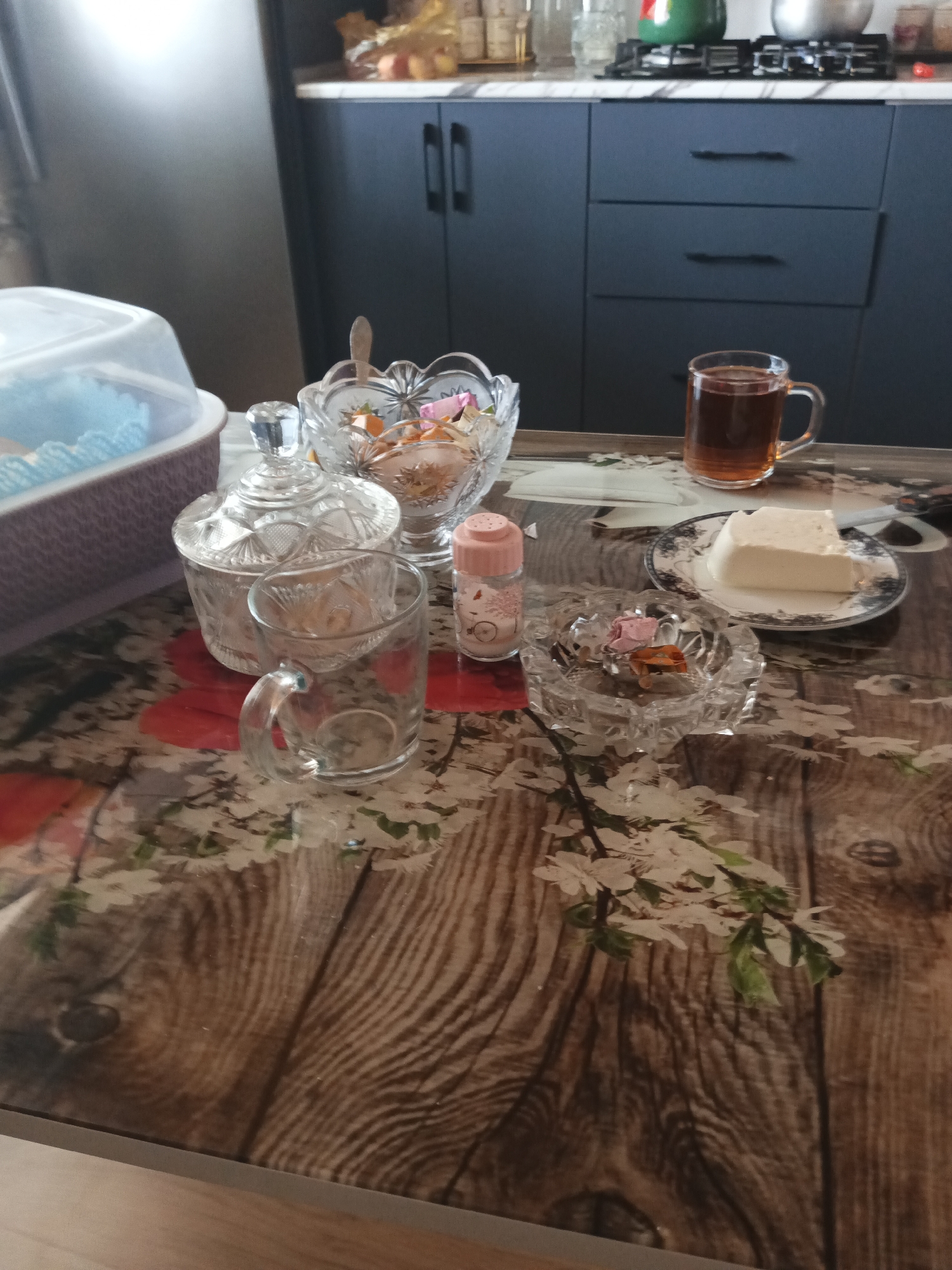 A close-up, eye-level shot depicts a wooden table with a floral and wood grain pattern. On the table, several items are arranged, suggesting a meal or gathering. To the left, a white plastic container with a clear lid holds blue cupcake liners, suggesting a dessert. Beside it sits a clear glass sugar bowl with a lid, containing a small spoon. A clear glass mug, empty, is placed in front of the sugar bowl. In the center, a decorative glass bowl holds candies and small colorful objects. To the right of the bowl, a small pink shaker, possibly for salt or sugar, stands. Further right, a clear glass ashtray contains a few pieces of colorful candy. Behind the ashtray, a slice of white cheese or cake sits on a decorative plate. A clear glass mug filled with amber-colored tea is positioned behind the plate. The background shows dark blue kitchen cabinets and a stovetop, indicating the location is a kitchen. The image is well-lit, with natural light casting soft shadows. The overall impression is one of a cozy domestic setting, possibly in Masazir, Azerbaijan, with remnants of a shared meal or treat.