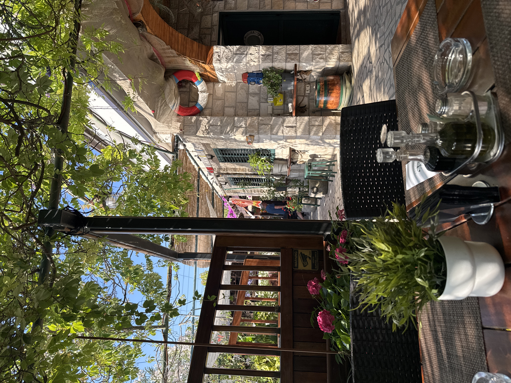 The image shows an outdoor seating area of a restaurant with a rustic, coastal theme. In the foreground, a table is set with a woven placemat, a glass ashtray, salt and pepper shakers, and two glass cruets containing dark liquid and green liquid, possibly balsamic vinegar and olive oil. A white potted plant with green foliage sits on the table.
In the mid-ground, a dark woven chair is visible. Behind the table, a wooden fence with vertical slats separates the seating area from a garden with vibrant pink hydrangeas. A "WARNING 24H VIDEO SURVEILLANCE" sign is attached to the fence. Beyond the fence, a pathway leads to a building with stone walls and arched entrances. Several potted plants and decorative items, including a life preserver and a barrel, are visible on and around the building. Sunlight casts long shadows across the scene, indicating it is late afternoon or early morning. The overall atmosphere suggests a relaxed, Mediterranean-style dining experience.