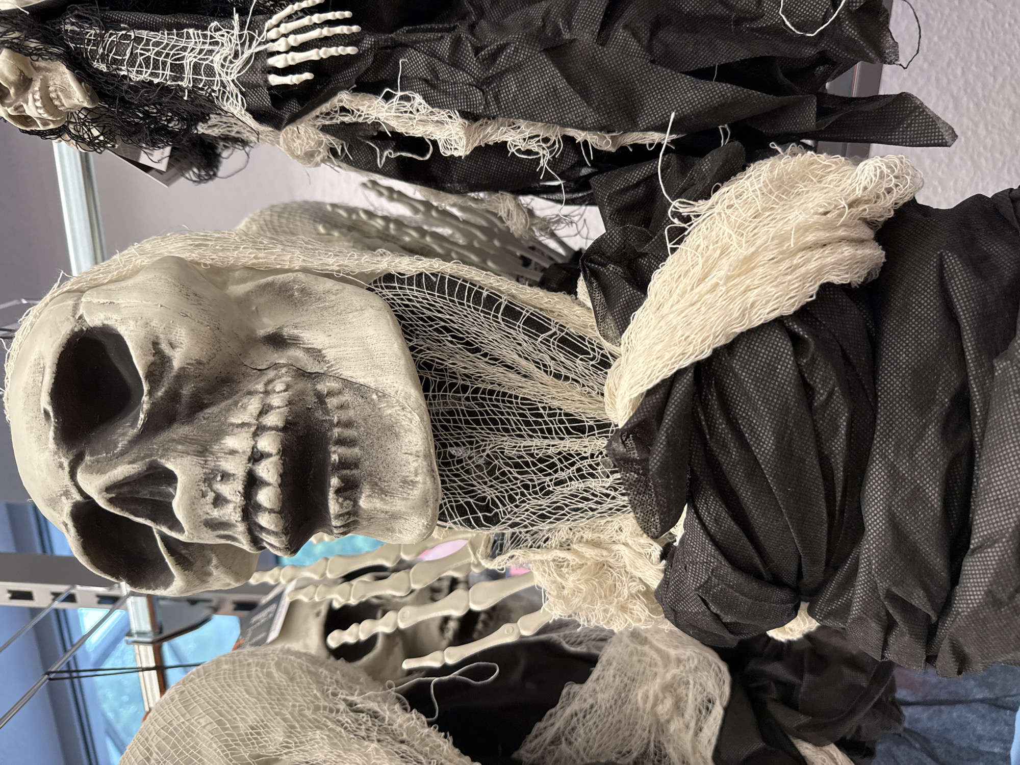 The image shows a close-up of several Halloween decorations, specifically skull figures, hanging from a display rack. The figures are adorned with tattered fabric and netting in white and black, giving them a spectral appearance. In the foreground, a large skull with visible teeth and eye sockets dominates the frame. Behind it, parts of other similar figures are visible, including skeletal hands and more fabric. The background reveals a slightly out-of-focus view of what appears to be a store interior, with shelving and possibly a window showing a glimpse of the outdoors. The overall scene is set in a retail environment, likely a store selling seasonal decorations, and could be located in Bad Lauterberg im Harz, Germany, where such items would be displayed for sale.