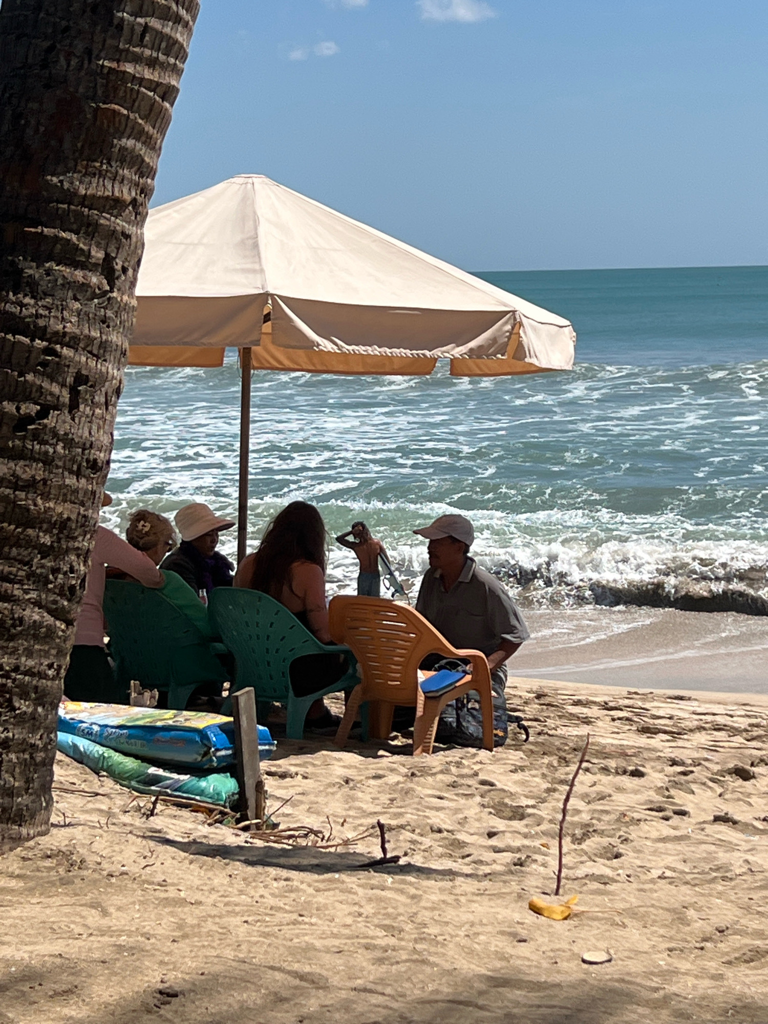 In Kuta, Indonesia, a group of people are gathered on a sandy beach under a large umbrella. The ocean waves are visible in the background, with a clear blue sky above. A palm tree trunk is in the foreground on the left, partially obscuring the view. In the center, several individuals are seated on chairs, facing each other. One person, a man wearing a cap and a dark shirt, is seated on an orange chair and looking to his left. Another man is standing in the water with a surfboard. Several beach mats are piled on the sand to the left of the seated group.