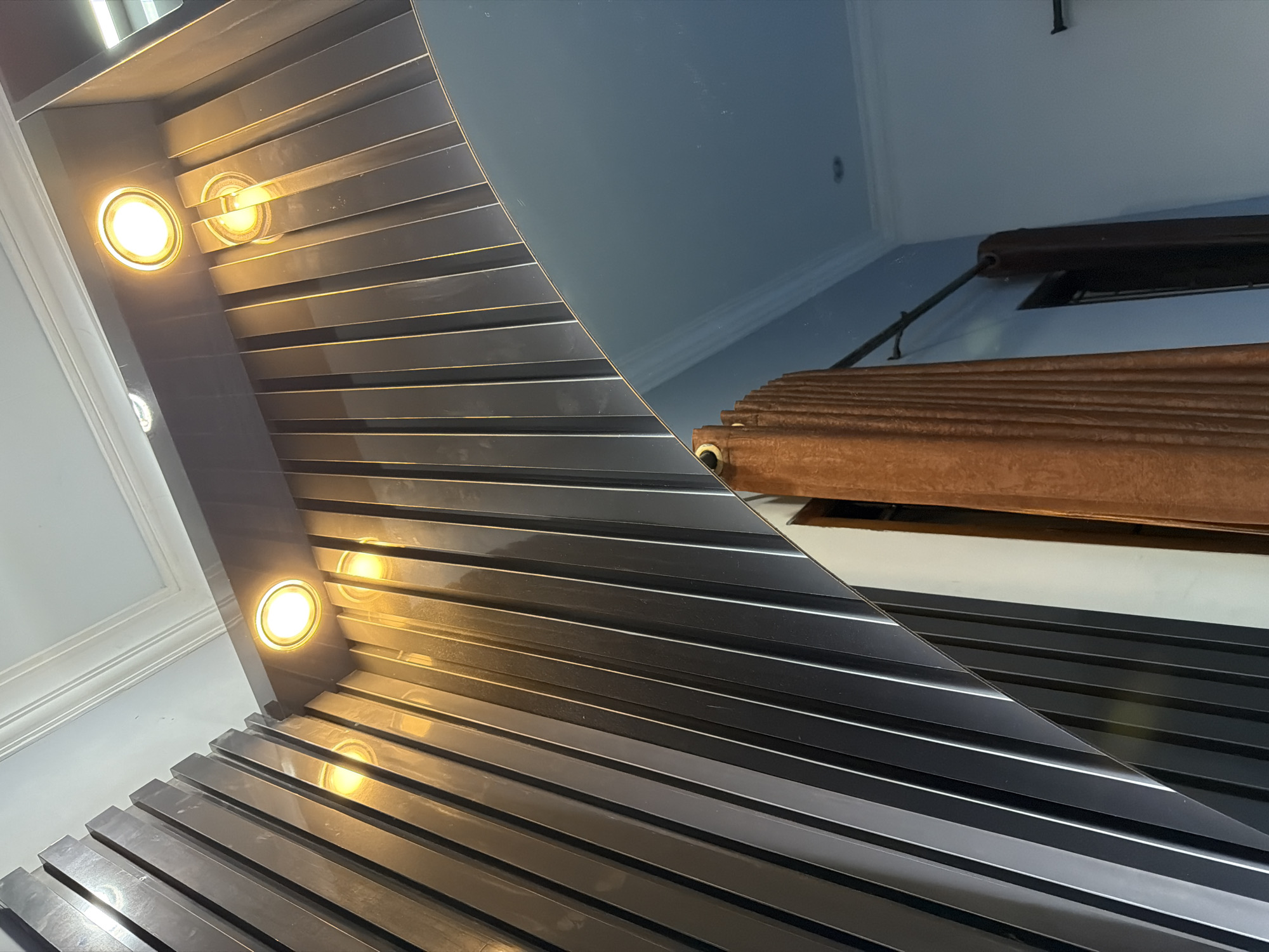 The image depicts an interior scene, possibly a hallway or a large room, featuring a modern design with reflective surfaces and lighting. The main element is a dark, ribbed structure that angles upwards and to the left. Three circular spotlights are embedded in the upper part of this structure, emitting a warm yellow light. To the right, a large, curved mirror reflects a portion of the room. In the reflection, a brown, pleated curtain is visible, hanging from a dark rod attached to a light blue wall. The ceiling is white with a decorative molding. The overall impression is one of contemporary interior design. The scene does not contain any people. Based on the available information, the location is identified as Wonoayu, Indonesia.