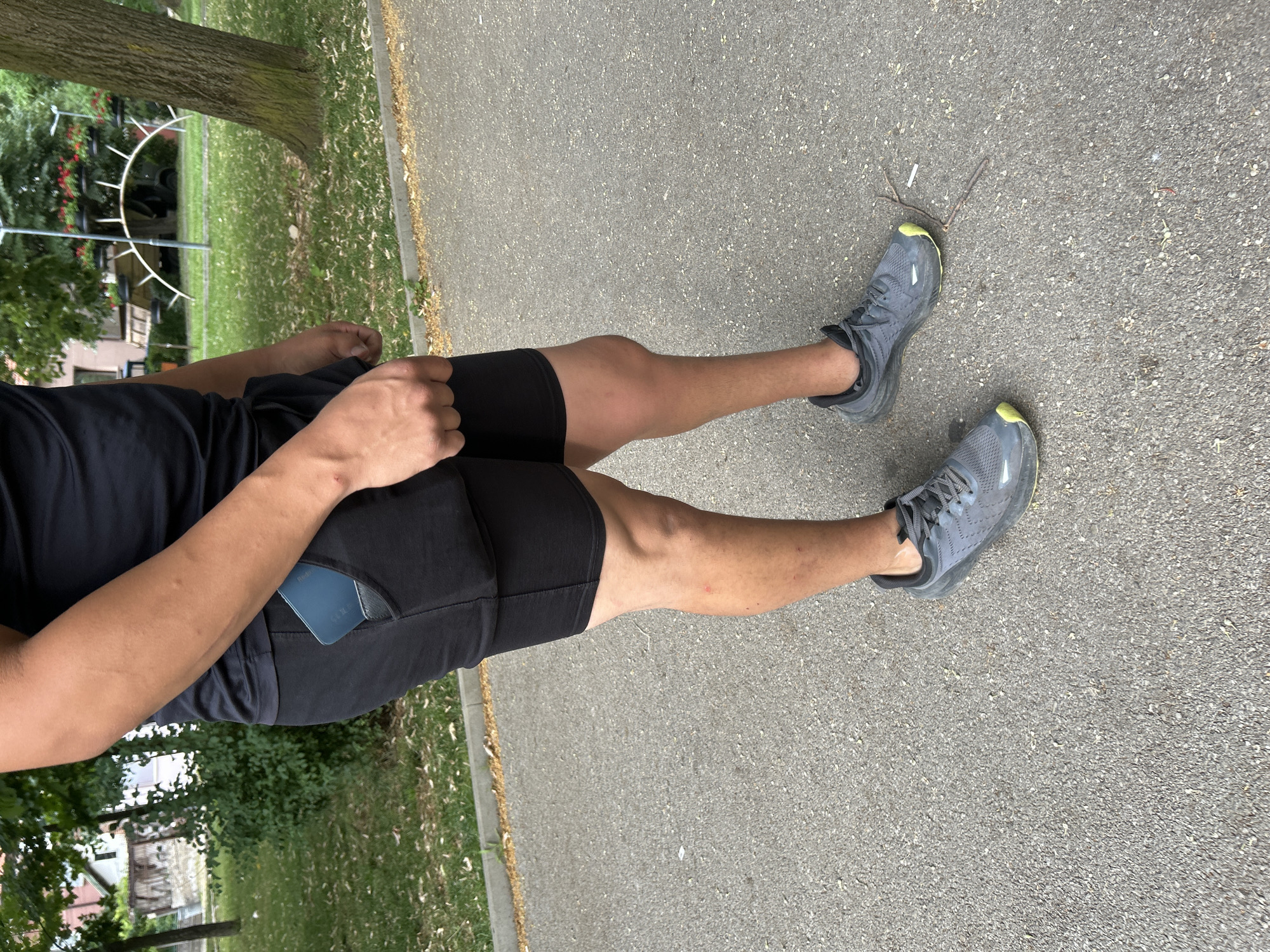 A person is standing on a paved walkway outdoors, with a grassy area and trees in the background. The person is wearing black athletic shorts with a built-in phone holder containing a blue smartphone, a black t-shirt, and grey athletic shoes with yellow accents. The person's left hand is clenched and resting near their shorts. The environment suggests a park or recreational area within the city of Vratsa, Bulgaria, given the presence of mature trees, manicured grass, and a building visible in the distance. The pavement is asphalt, showing some wear and small debris.