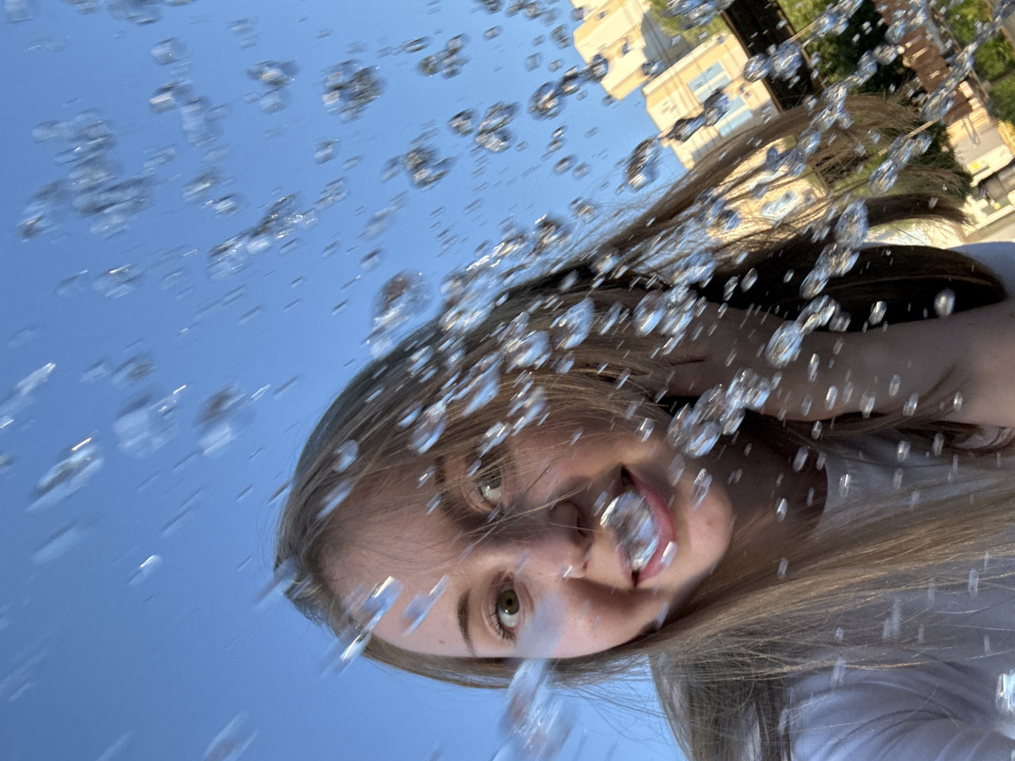 In this low-angle, close-up, outdoor, eye-level shot, a young Caucasian woman with long, straight blonde hair is captured looking directly at the camera with a wide smile and slightly widened eyes. She is wearing a white t-shirt. The foreground is obscured by a cascade of water droplets or bubbles, some of which are in sharp focus, while others are blurred. The woman's face is partially framed by her hair, which is also affected by the water. In the background, out of focus, a building with light-colored walls and windows is visible, along with some greenery. The sky above is a clear, bright blue. The overall impression is one of playful interaction with water on a sunny day.