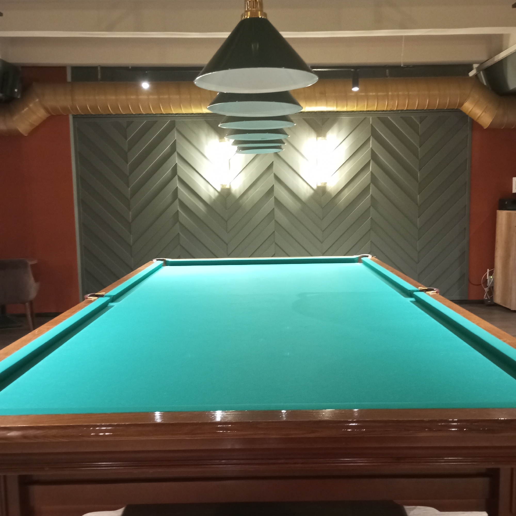 This is a medium-shot, eye-level, interior photograph of a pool table in a dimly lit room. The pool table, covered with a vibrant green felt, dominates the foreground and middle ground. It has a polished wooden frame with brass-lined pockets. In the background, a grey, chevron-patterned wall is illuminated by two square wall sconces that emit a warm light. Above the wall, a large, yellow, corrugated ventilation duct runs horizontally. Hanging from the ceiling are several dark green pendant lights, arranged in a descending order. The lighting casts shadows, creating a moody atmosphere. To the left of the pool table, a portion of an orange wall and a blurred brown chair are visible. To the right, a wooden cabinet with visible wiring and a black speaker is present. The image does not provide any specific identifying information about the location within Izhevsk, Russia.