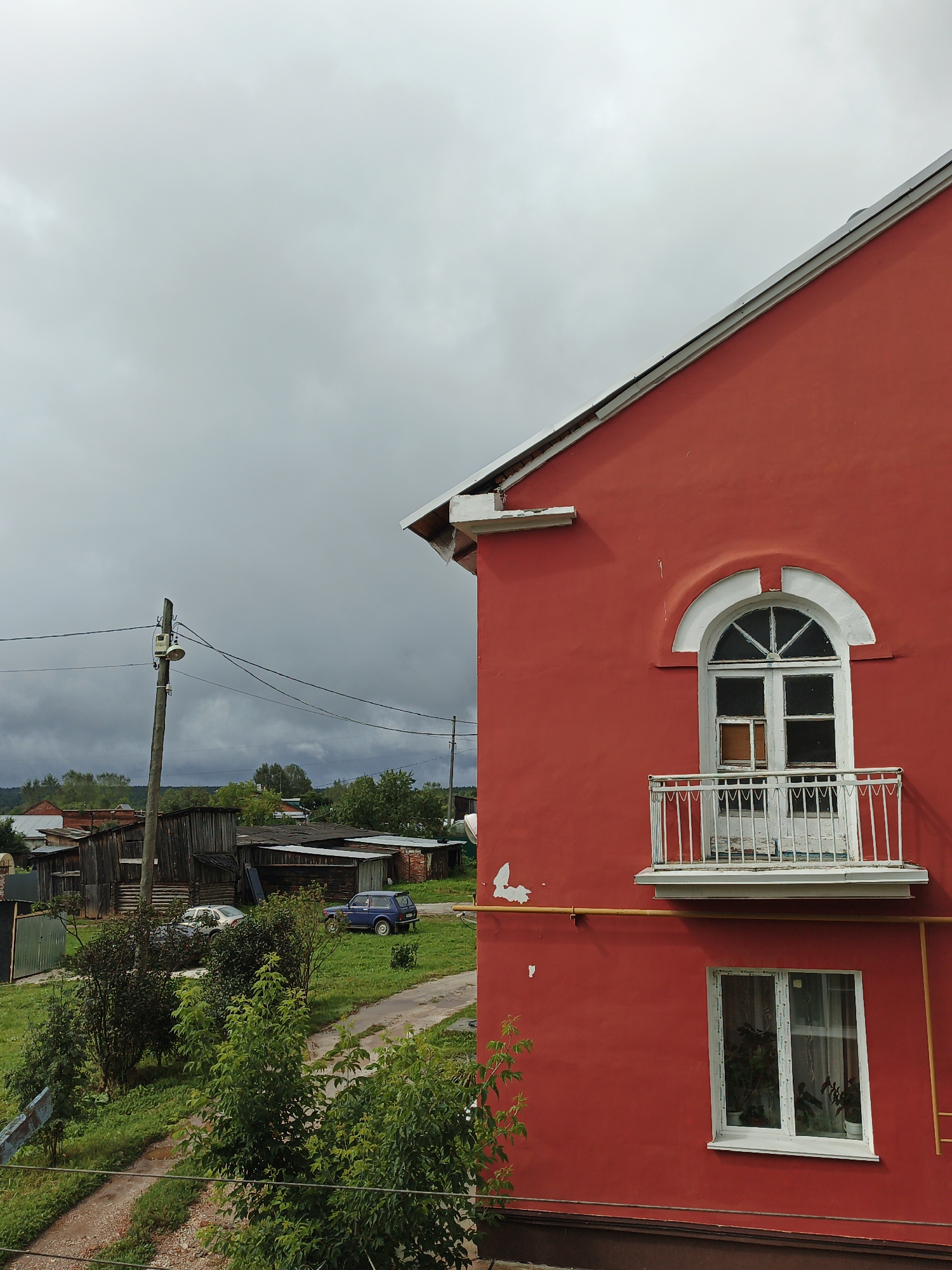 A bright red, two-story building with a white arched window and a small white balcony on the upper floor, and a white-framed window with plants on the lower floor, dominates the right side of the frame. A yellow pipe runs horizontally across the red facade. The building is situated in a rural setting, likely Medvedyovka, Russia. The left side of the image shows a cloudy, grey sky with dark clouds. Below the sky, a wooden utility pole with a light fixture and wires stands tall. In the middle ground, there are several rustic wooden buildings, some with corrugated metal roofs, and a dirt path. A blue car is parked on the path, and a white car is visible behind it. Lush green trees and bushes are scattered throughout the scene, with a dirt road leading into the foreground. The overall impression is a somewhat overcast day in a village or rural community. There are no people visible in the image.