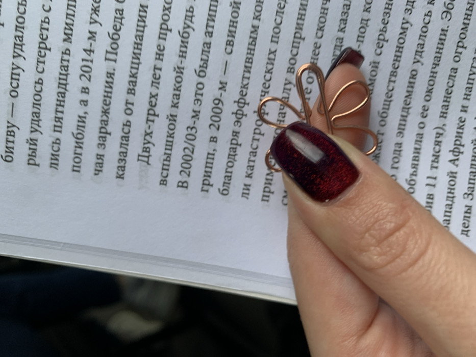 The image presents a close-up view of a human left hand holding a decorative paperclip on a printed page. The hand exhibits light skin with a manicured index finger adorned with dark red, shimmering nail polish. The thumb is partially visible, bracing the object. The paperclip is crafted from thin, shiny copper-colored wire, formed into a four-petal flower shape, and is clipped onto a white page of a book or document. The page displays black Cyrillic text, which discusses historical and epidemiological topics including smallpox, vaccinations, flu outbreaks (specifically atypical flu in 2002/03 and swine flu in 2009), and the Ebola epidemic. The immediate background to the upper left is dark and out of focus, while the main background is the text-filled page. The specific location within the city of Kozyn, Ukraine, is not discernible from the visual information provided in the image.